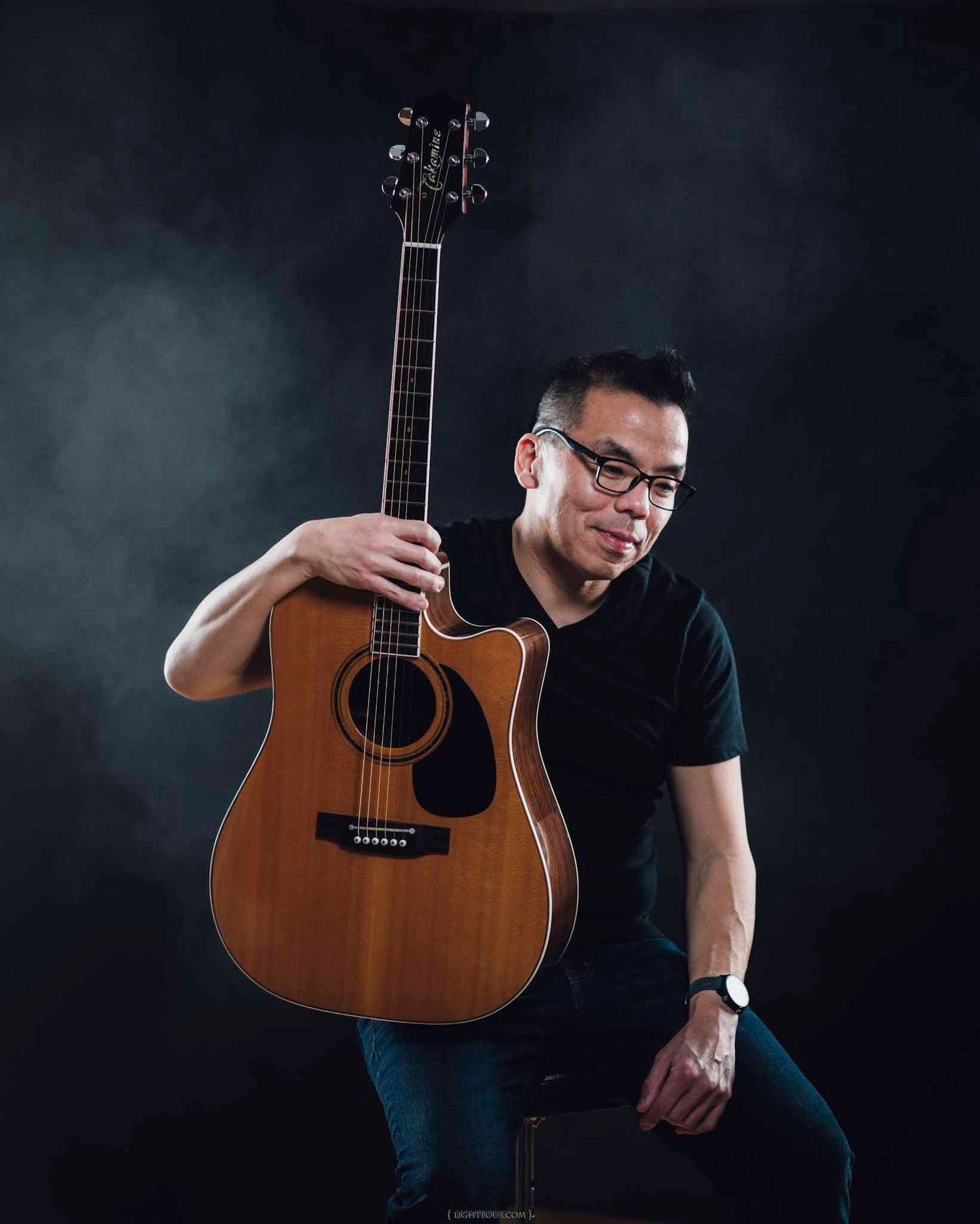 Man in black shirt holding acoustic guitar poses confidently against dark smoky background for professional music portrait.