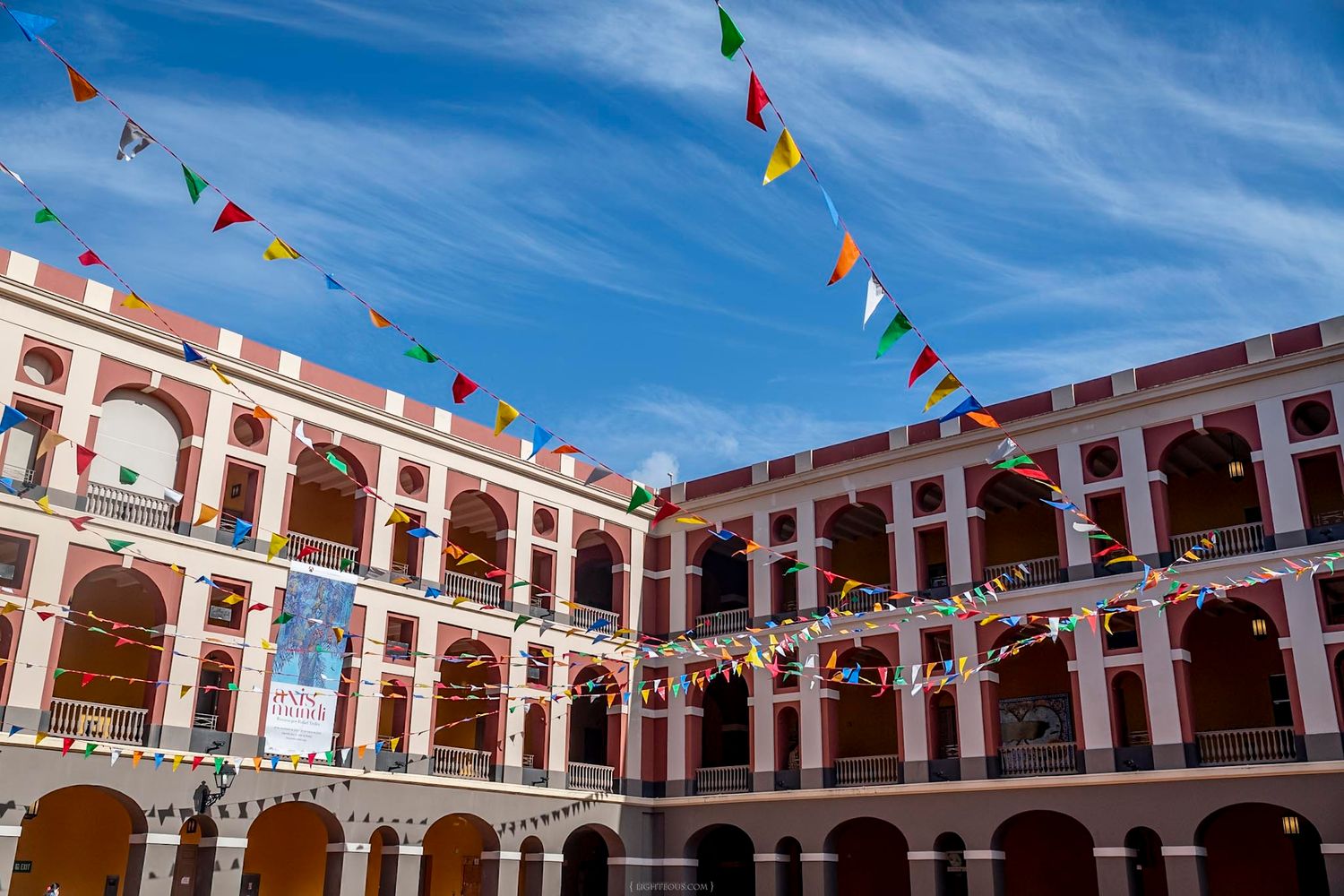 Colorful bunting flags decorate the courtyard of a historic red and white colonial building under blue sky.