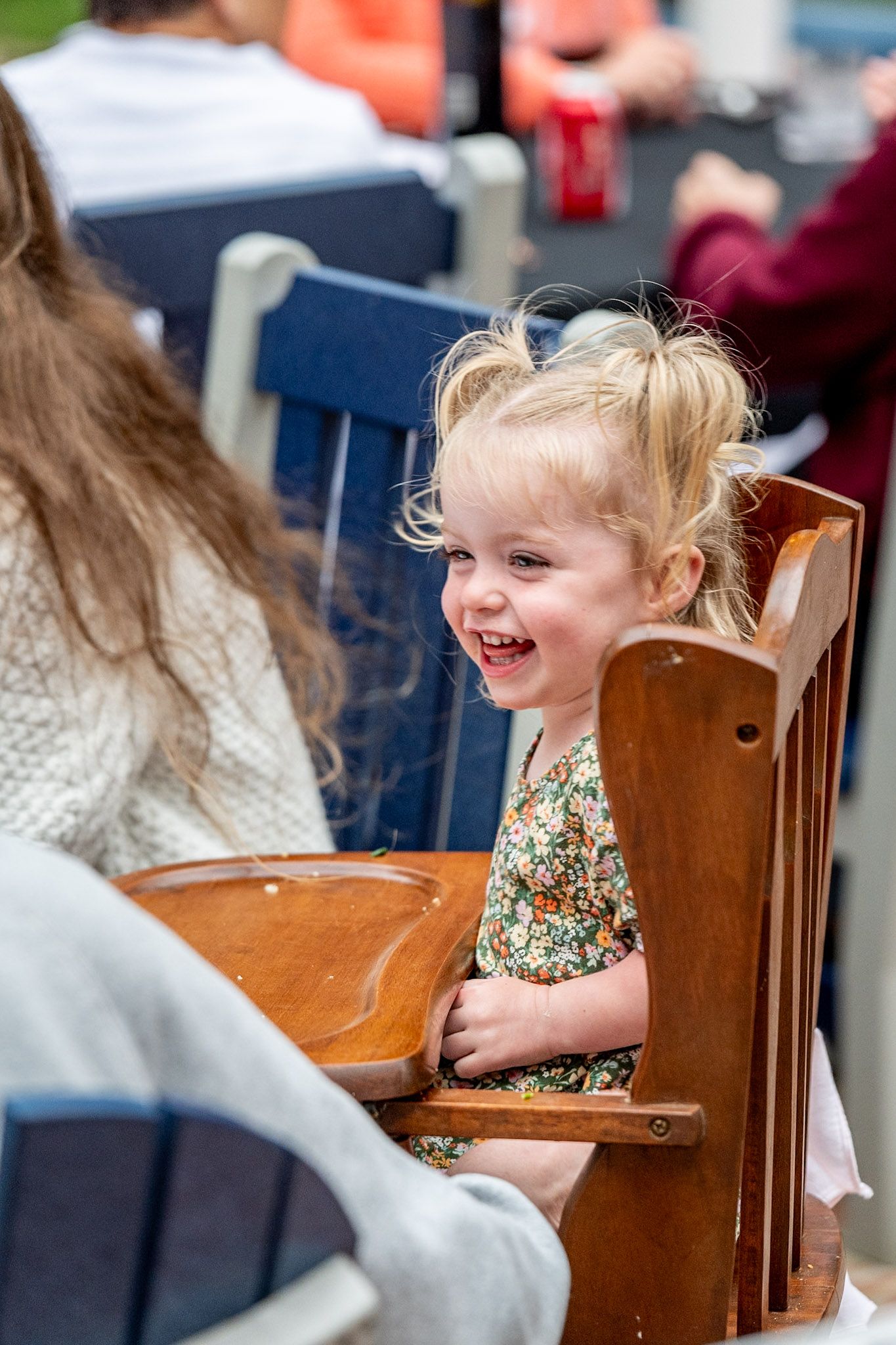 A child sits in a wooden chair at a table during an outdoor gathering.
