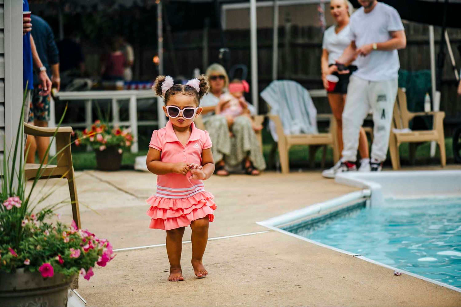 Toddler girl in pink dress and sunglasses standing by a pool at a backyard party.