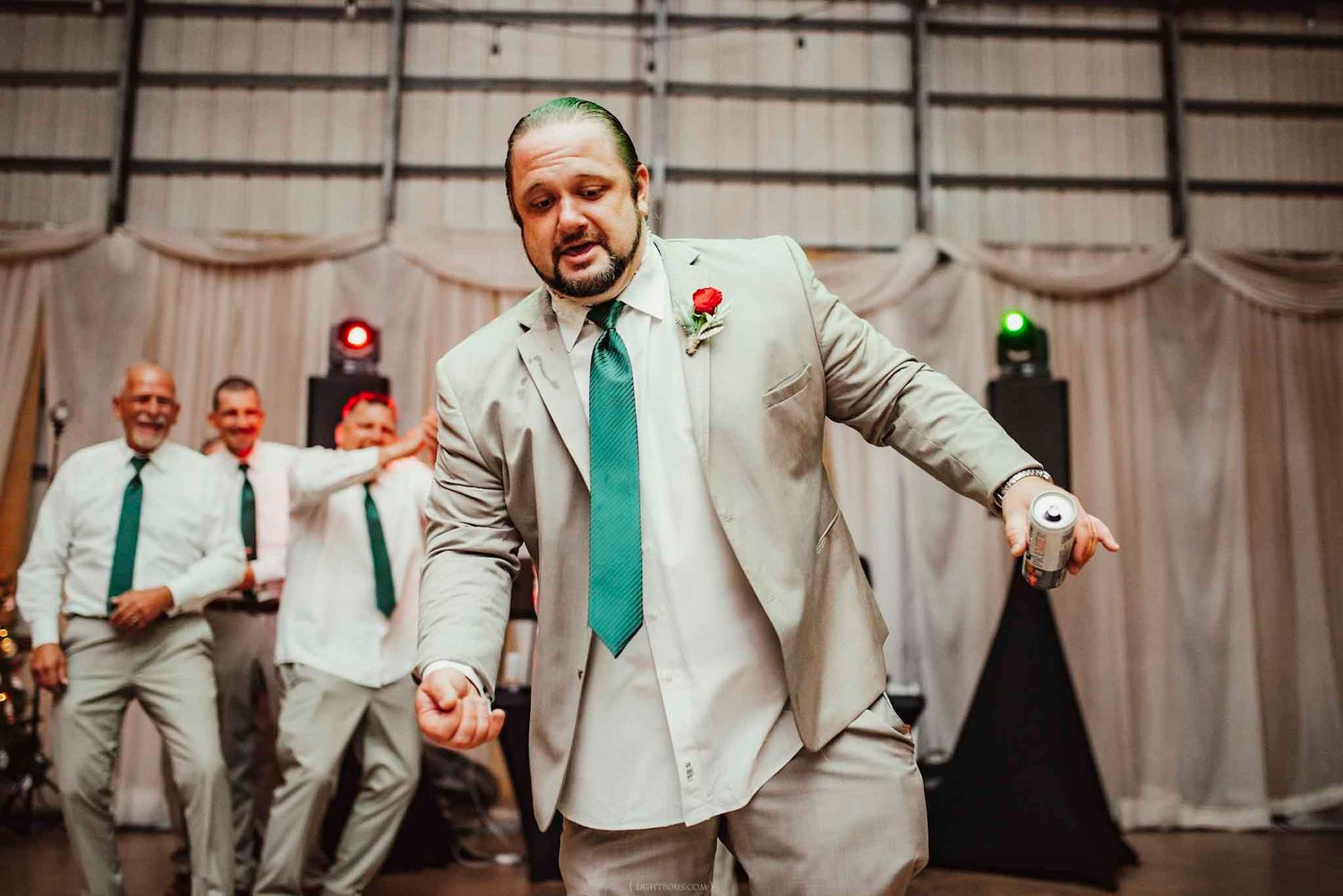 Groom dancing energetically at wedding reception in gray suit with teal tie, groomsmen celebrating behind him.
