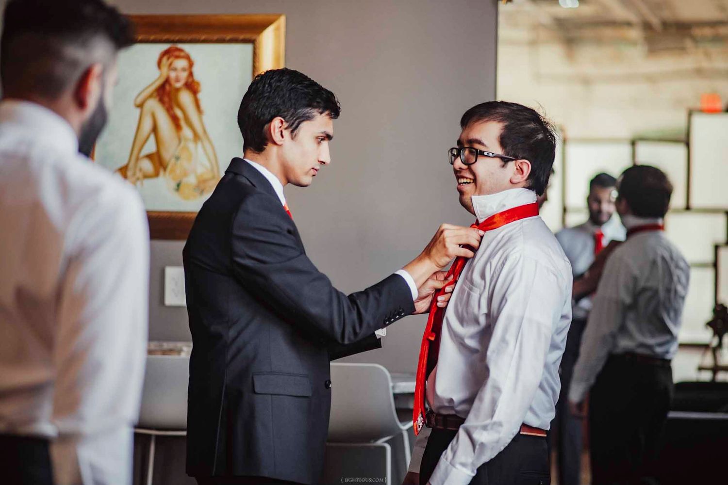 Two men in formal attire with red ties, one helping the other straighten his tie before an event.
