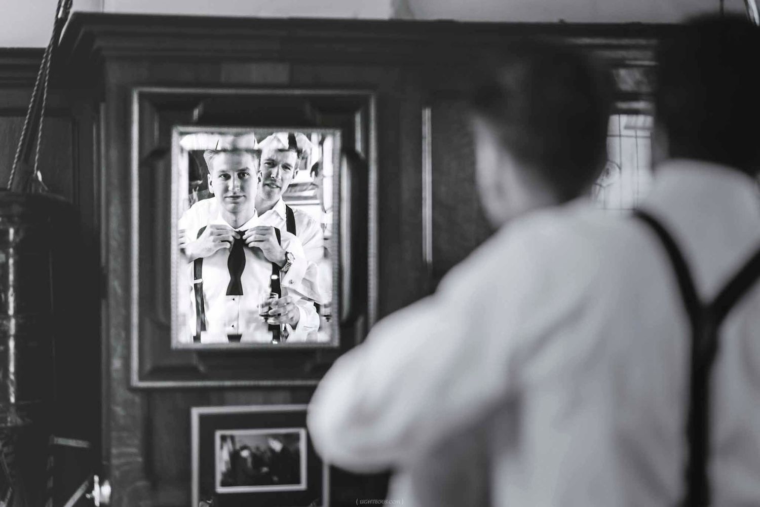 Black and white photo of a groom adjusting his suspenders while looking at his reflection in a mirror.