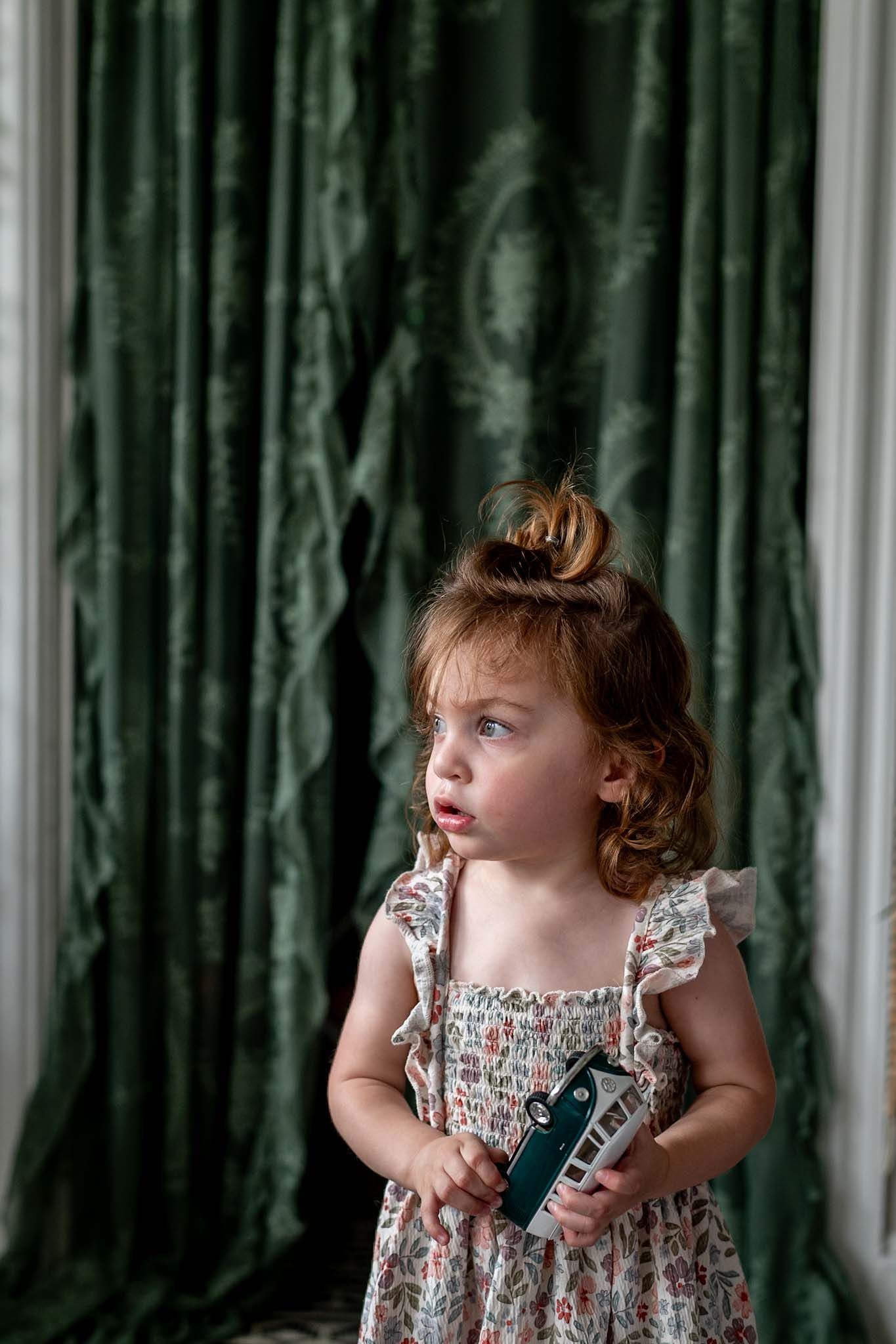Toddler girl in floral dress holding toy while standing in front of green velvet curtains.