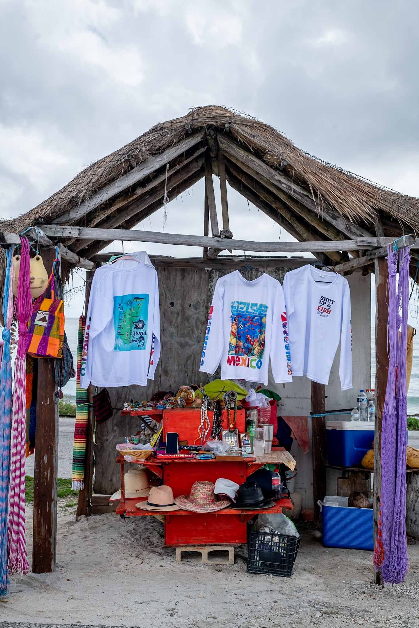 Beach vendor stall with thatched roof displaying colorful t-shirts and merchandise for sale on a cloudy day in Cozumel, Mexico.