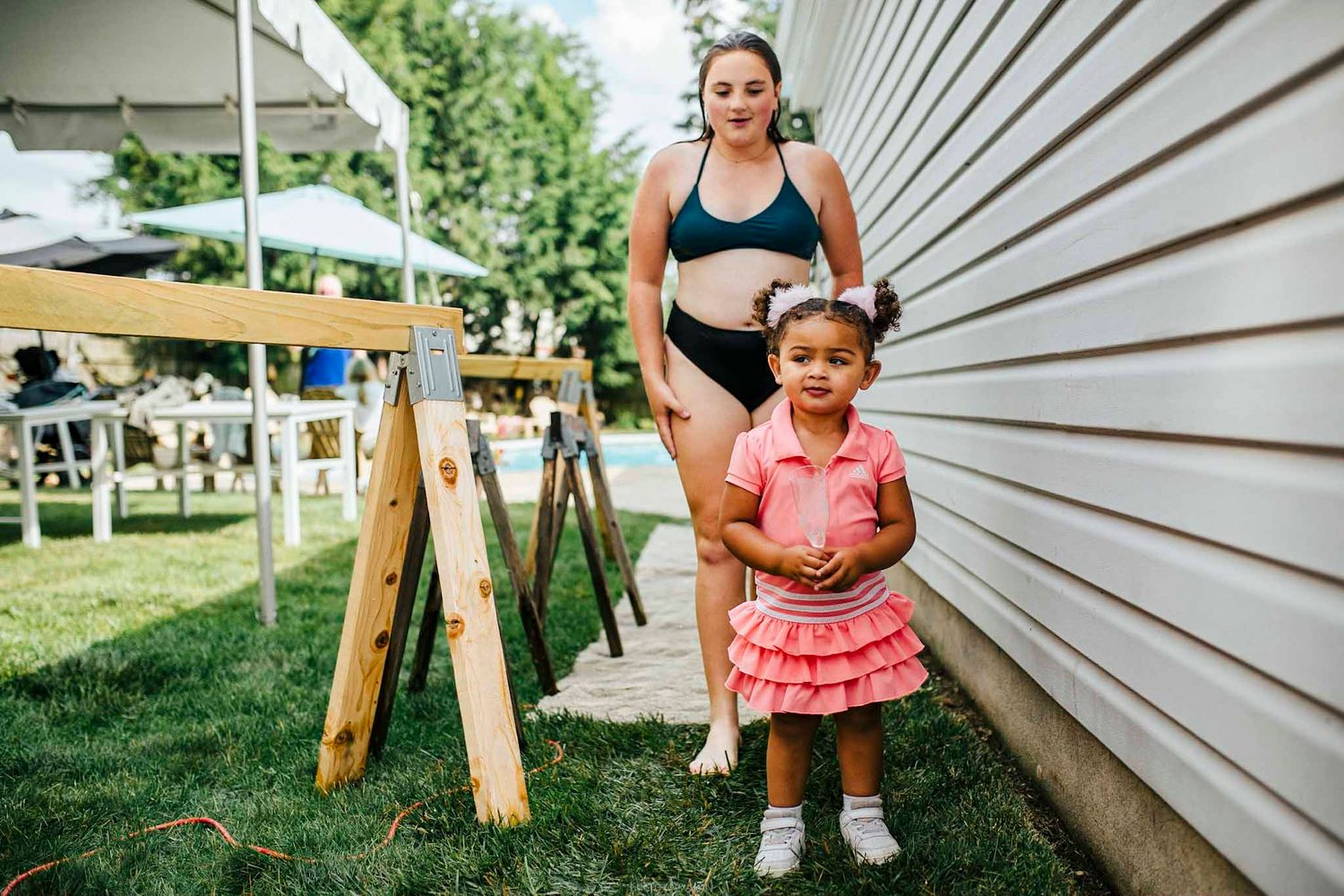 Woman in swimwear stands with young girl in pink dress outdoors near wooden boards and green lawn.