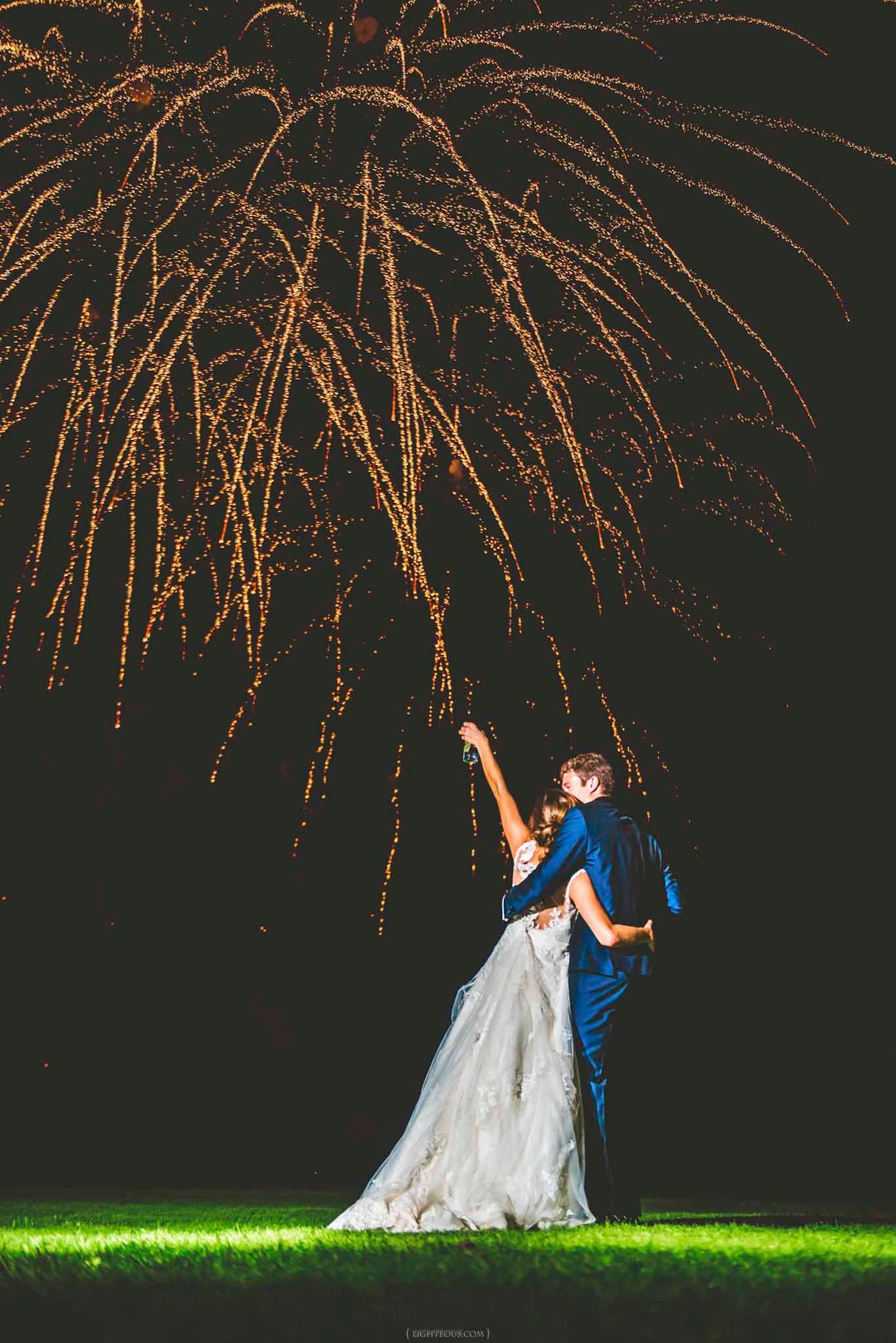 Bride and groom embrace on grass watching stunning golden fireworks light up the night sky at their wedding celebration.