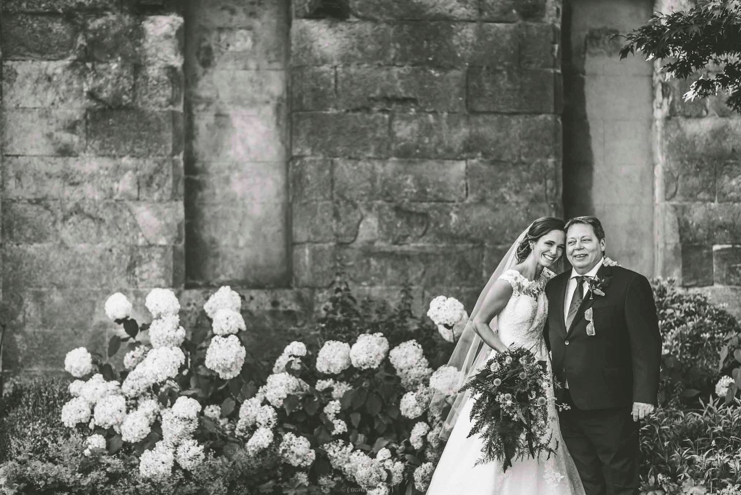 Black and white wedding photo of bride and groom posing by stone wall with blooming white hydrangeas.