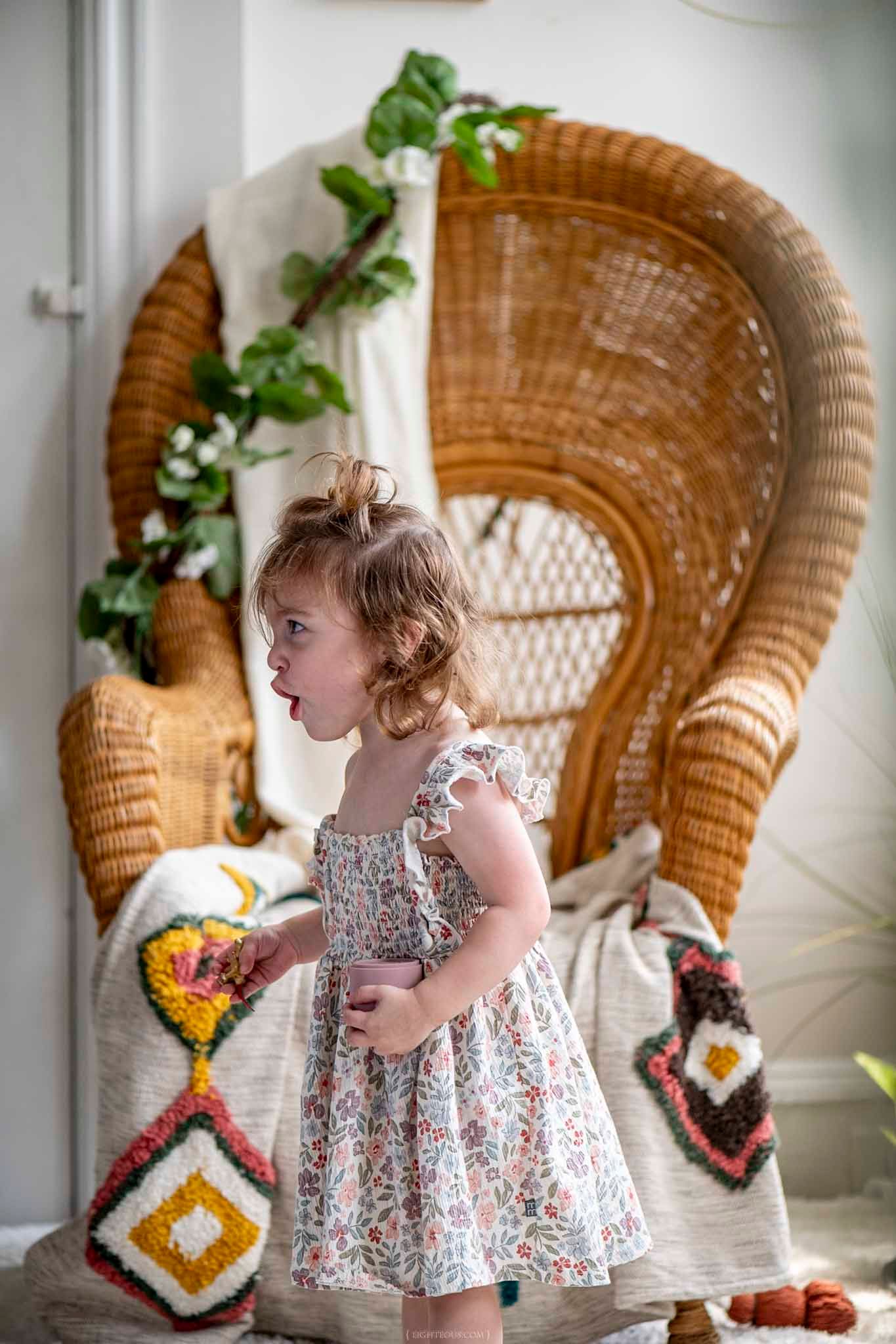 A young girl in a floral dress stands beside a large wicker peacock chair decorated with greenery and crochet pillows.