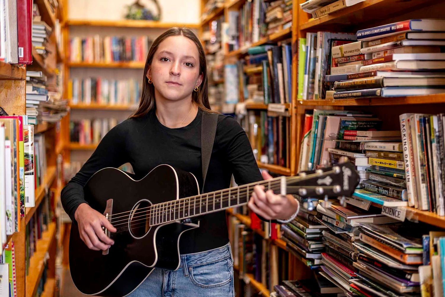 Young woman playing acoustic guitar among colorful bookshelves in a cozy bookstore setting.