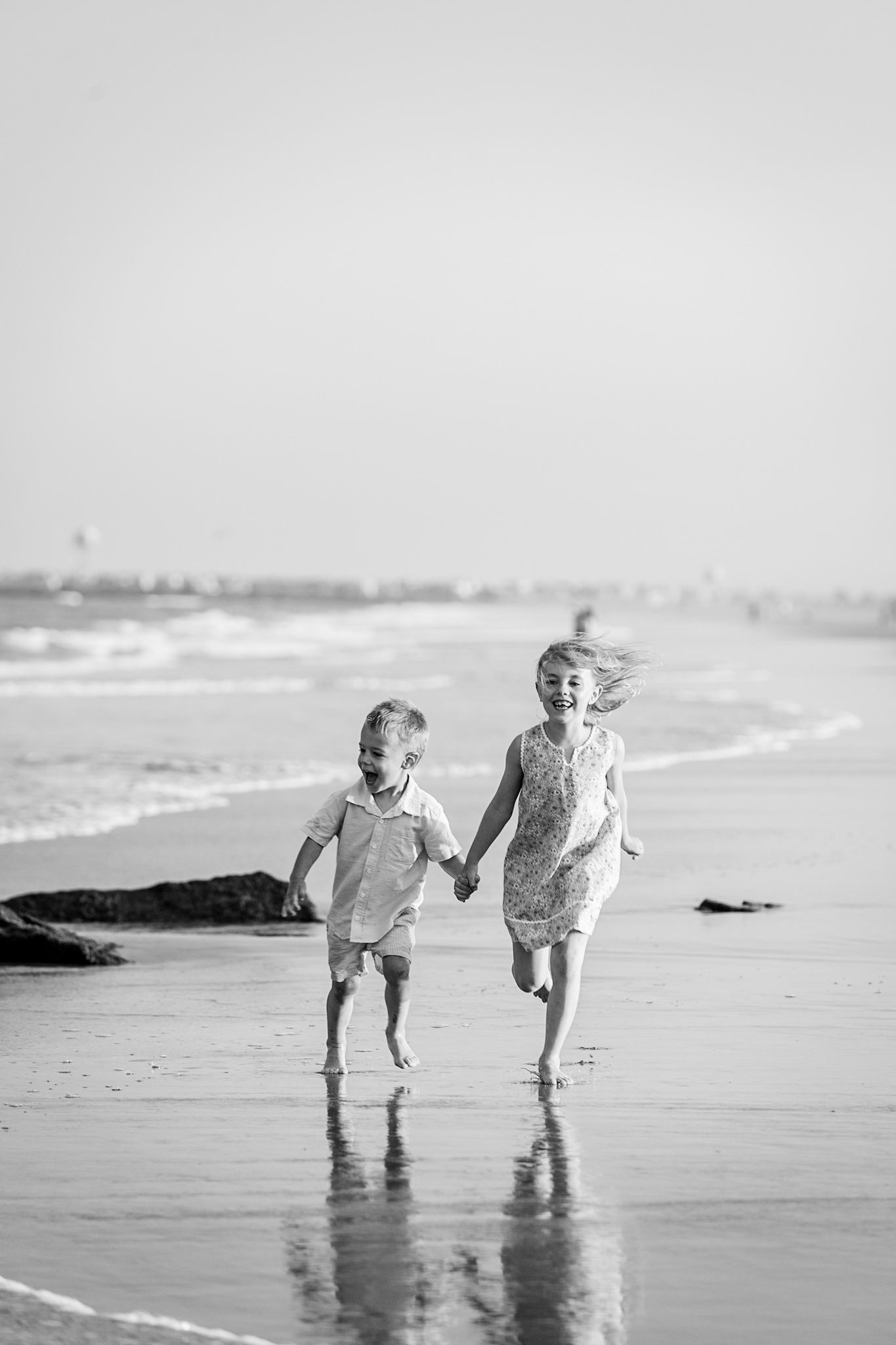 Two children holding hands and running joyfully along a sandy beach shore in a black and white photo.