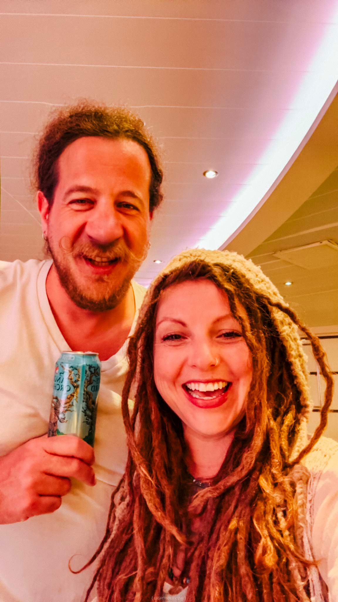 Smiling couple with dreadlocks holding a craft beer can indoors, enjoying a casual moment together.