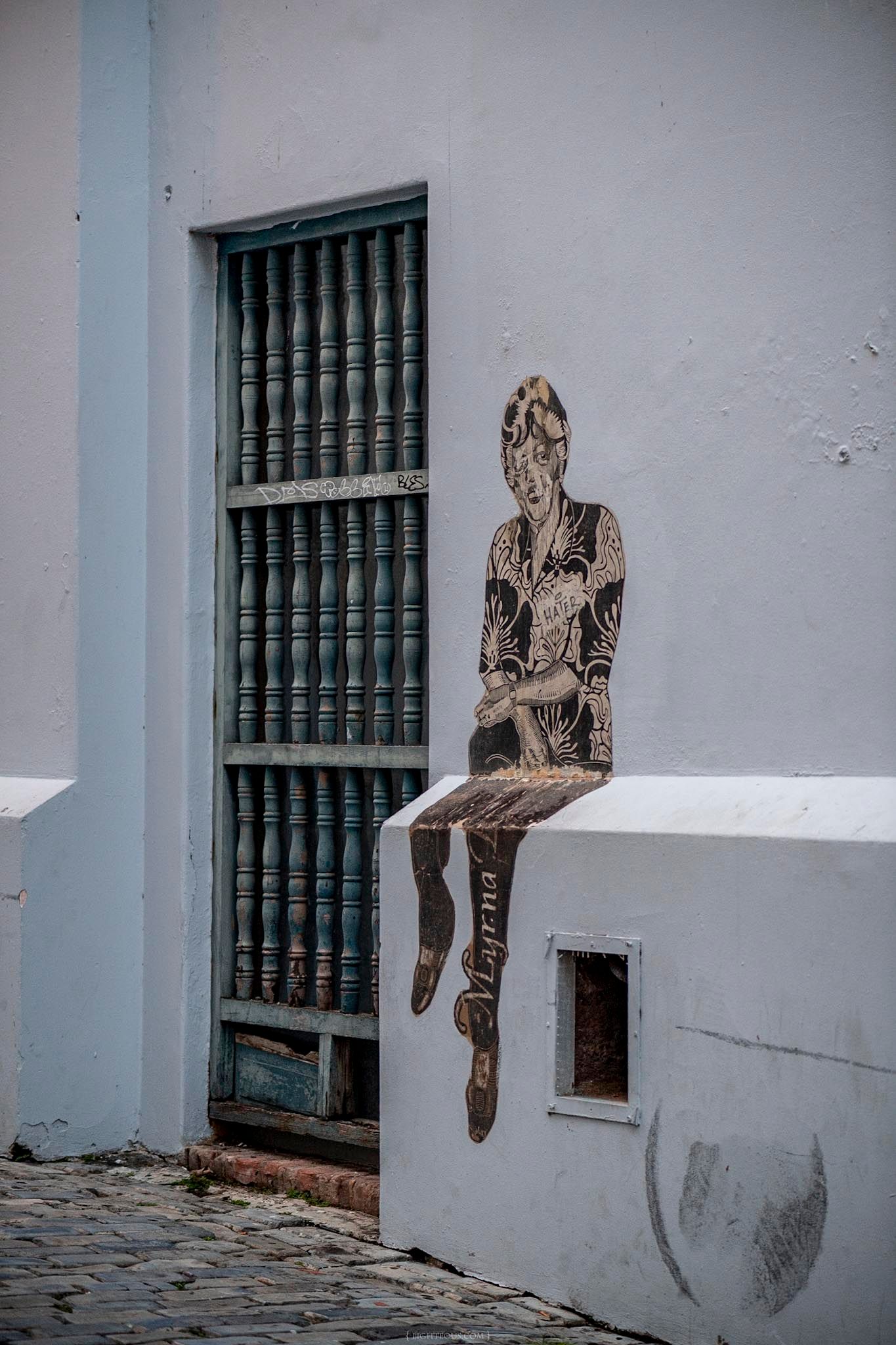 Street art mural of a tattooed figure sitting on a white wall ledge beside a barred window on a cobblestone street.