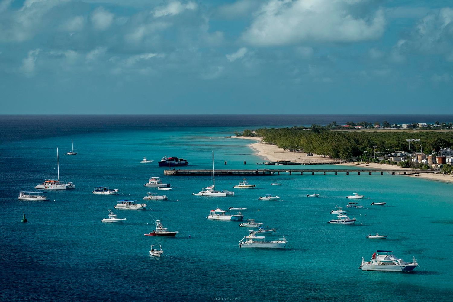 Aerial view of turquoise Caribbean harbor with multiple yachts and boats anchored near a sandy tropical beach.