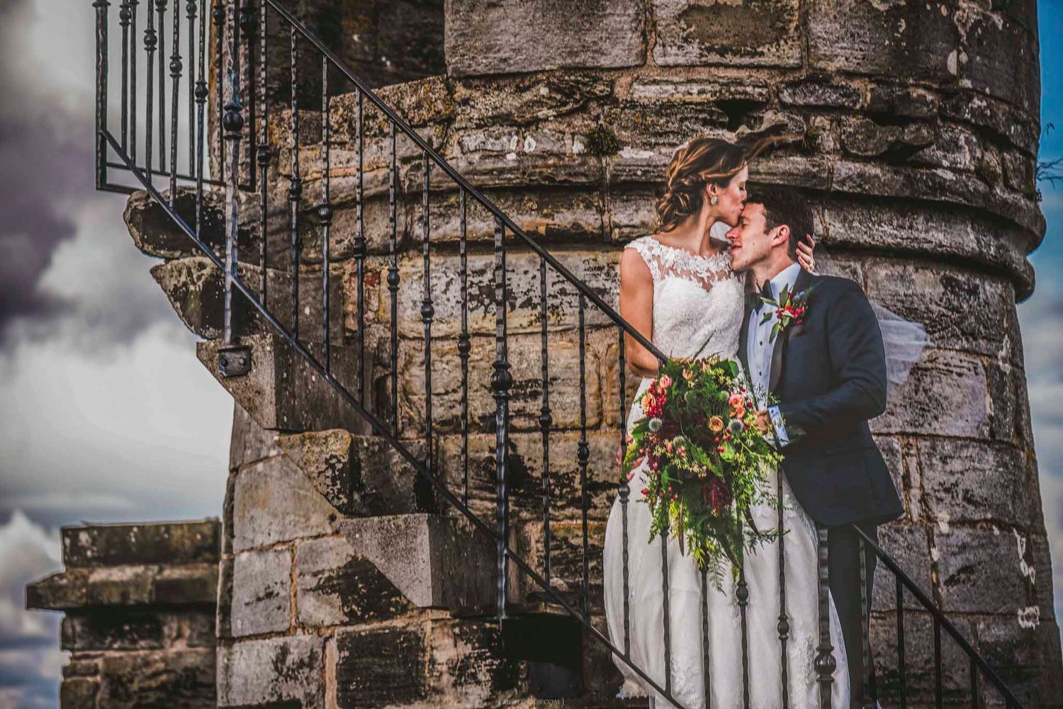 Bride and groom sharing a romantic moment on ancient stone spiral staircase with colorful bridal bouquet.