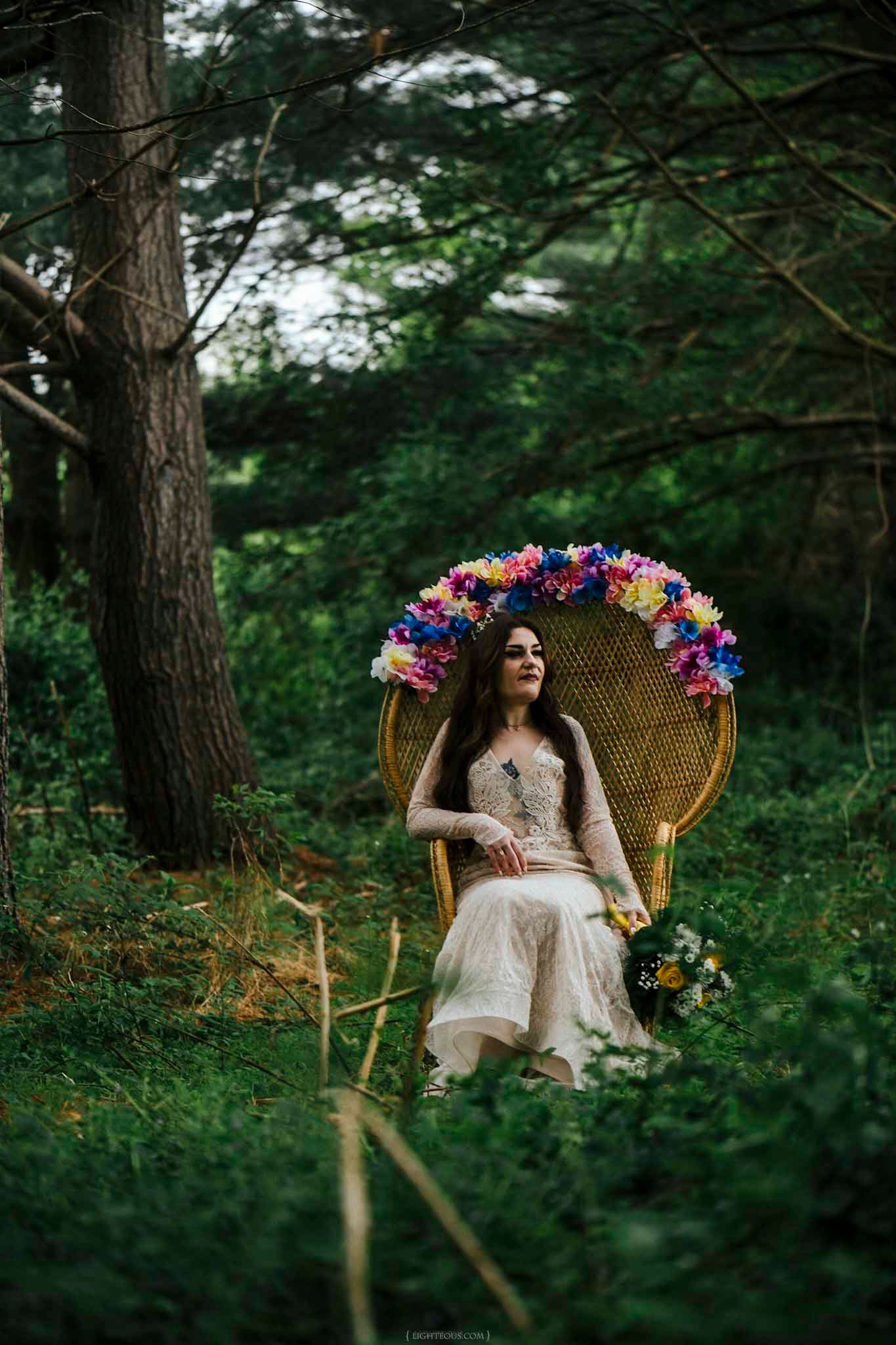 Bride in white gown sits in wicker peacock chair adorned with colorful flowers, surrounded by lush green forest.