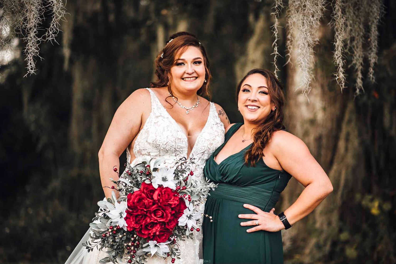 Bride in lace gown holding red bouquet poses with bridesmaid in green dress under Spanish moss-draped trees.