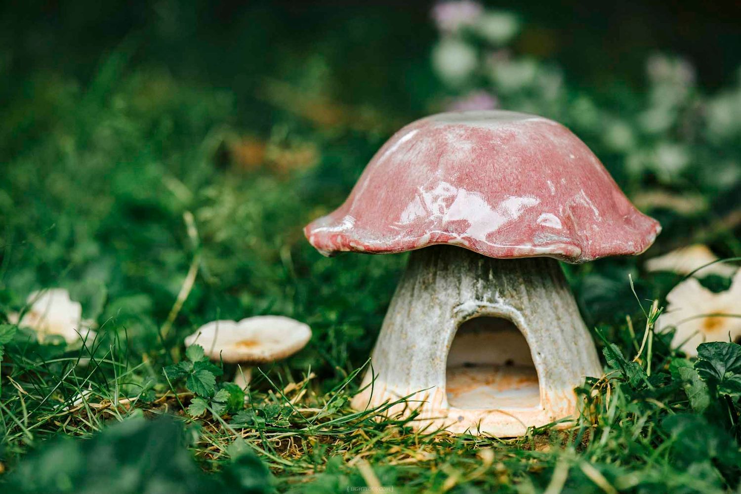 Ceramic mushroom house figurine with pink cap sitting in green grass surrounded by small white mushrooms.