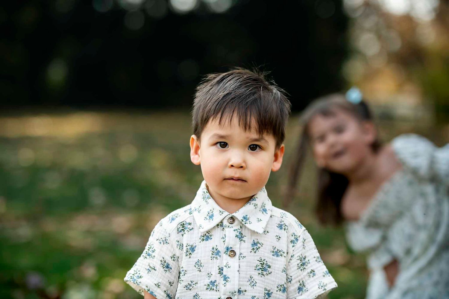 Young boy in floral shirt poses outdoors with girl in background at a park during golden hour.
