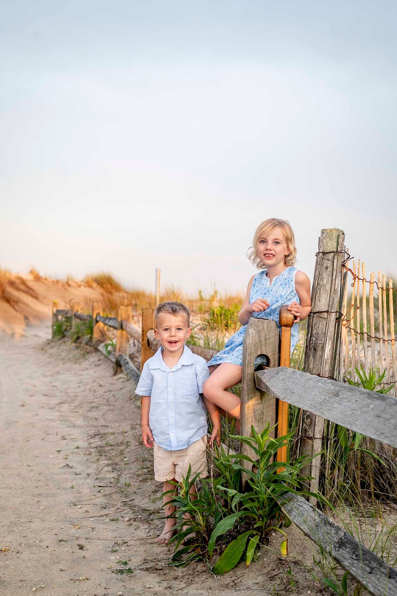 Two young children pose by a rustic wooden beach fence on a sandy path, boy in blue shirt and girl in floral romper.