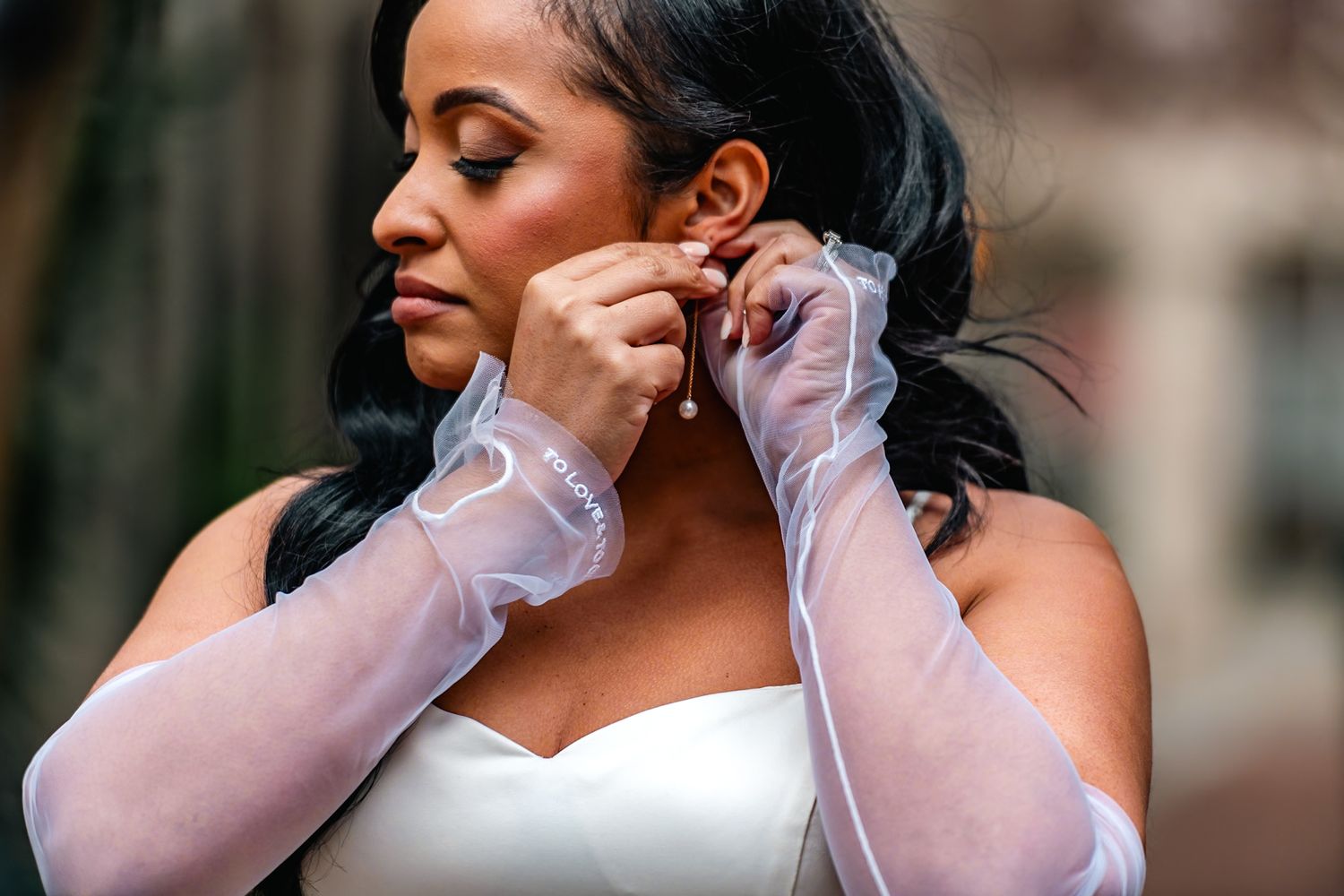 Bride in white strapless gown and sheer gloves putting on earrings outdoors on her wedding day.