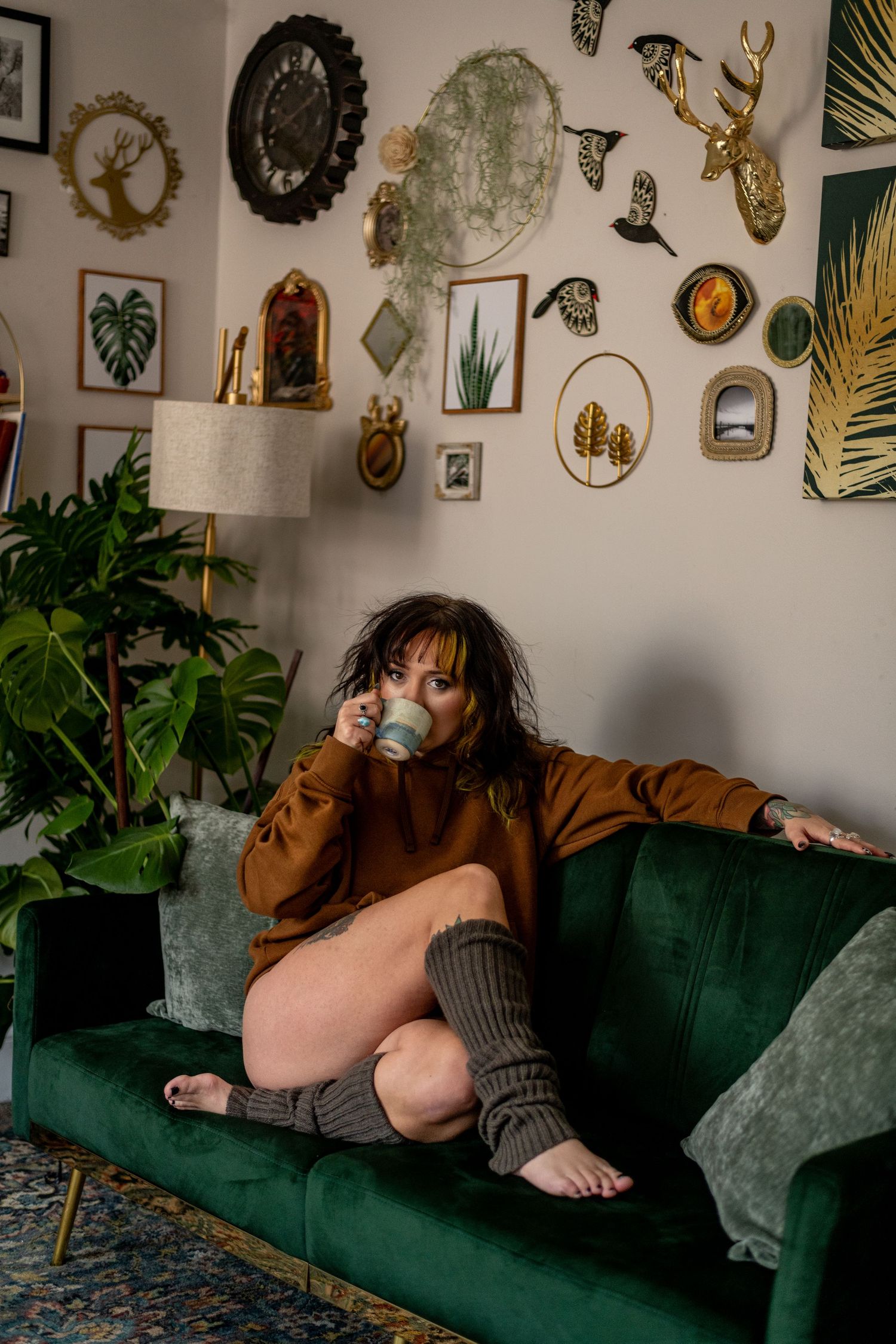 Woman in brown sweater and leg warmers drinking from a mug on a green velvet sofa.