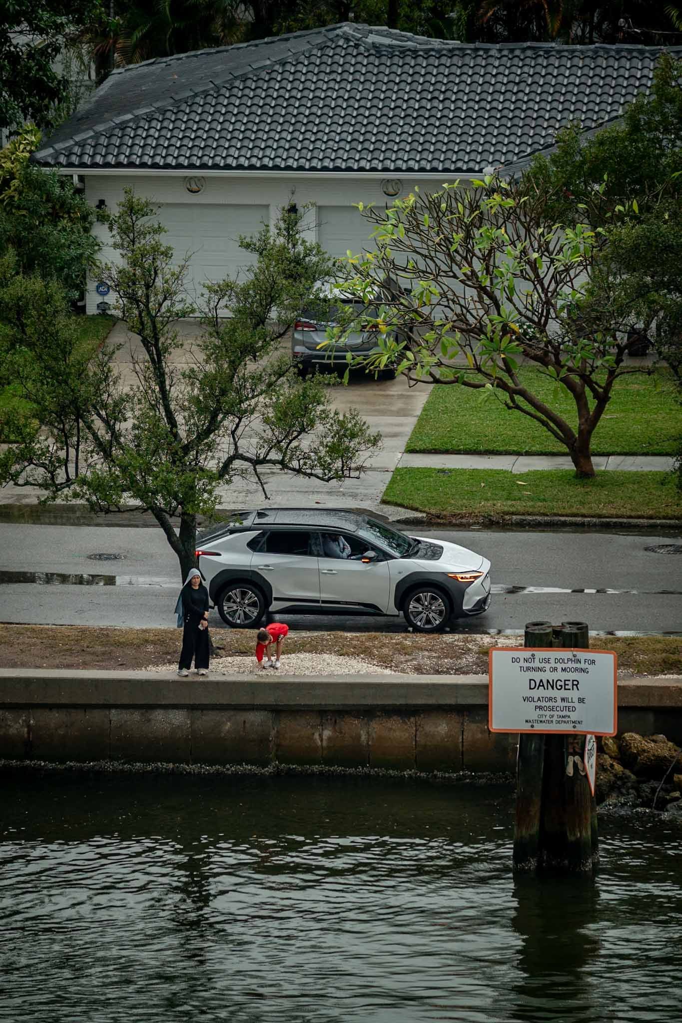 Warning sign posted along a waterfront walkway with parked cars and trees in the background.