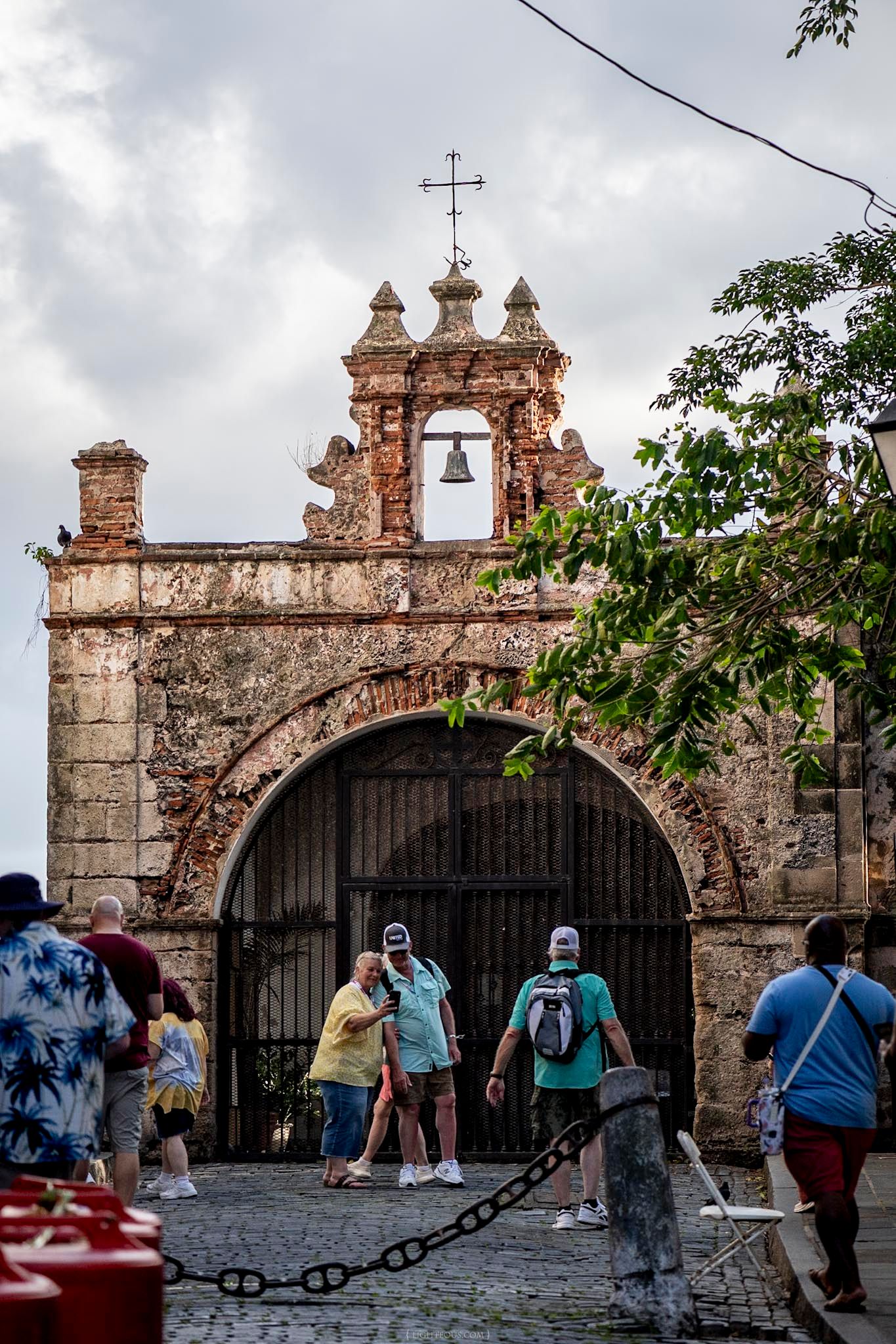 Tourists explore a historic stone archway with bell tower and cross in Old San Juan, Puerto Rico.