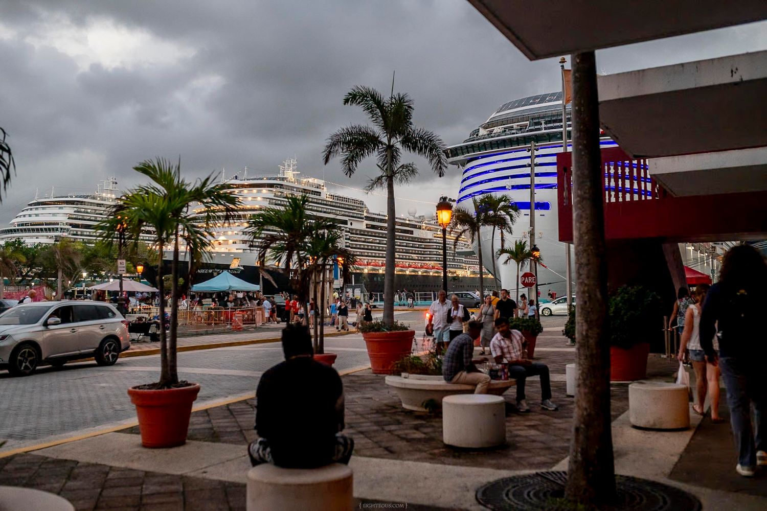 Cruise ship docked at a tropical port with palm trees, overcast skies, and people relaxing near a waterfront plaza.