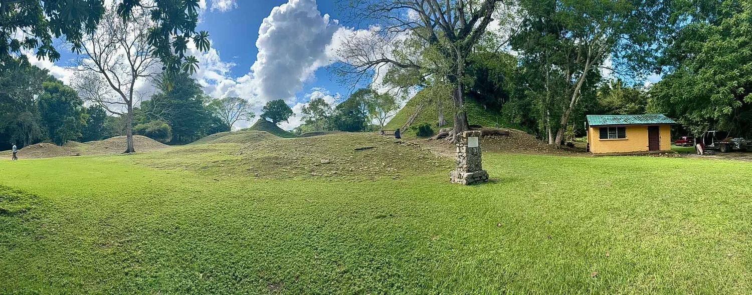 Panoramic view of a green lawn with trees and a small yellow building under puffy white clouds in a blue sky.
