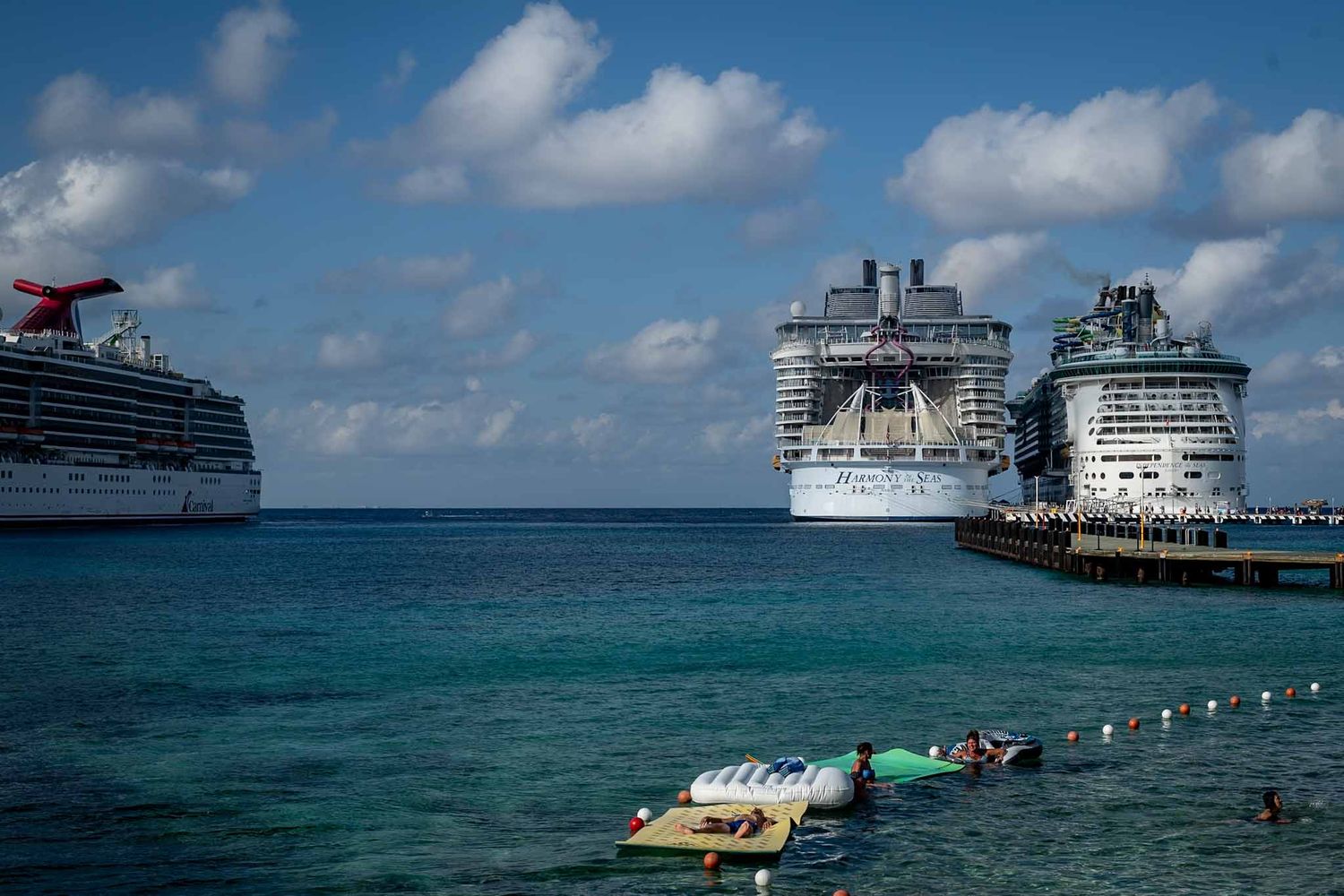 Cruise ships docked at a tropical Caribbean port with turquoise waters and floating swim platforms visible.