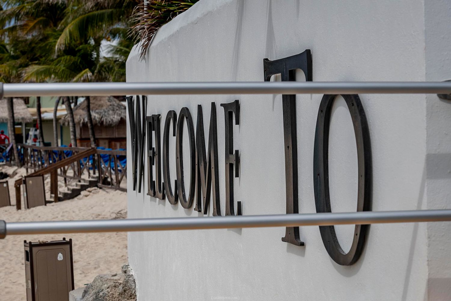 Welcome To sign on a white wall at a tropical beach resort with palm trees and lounge chairs.