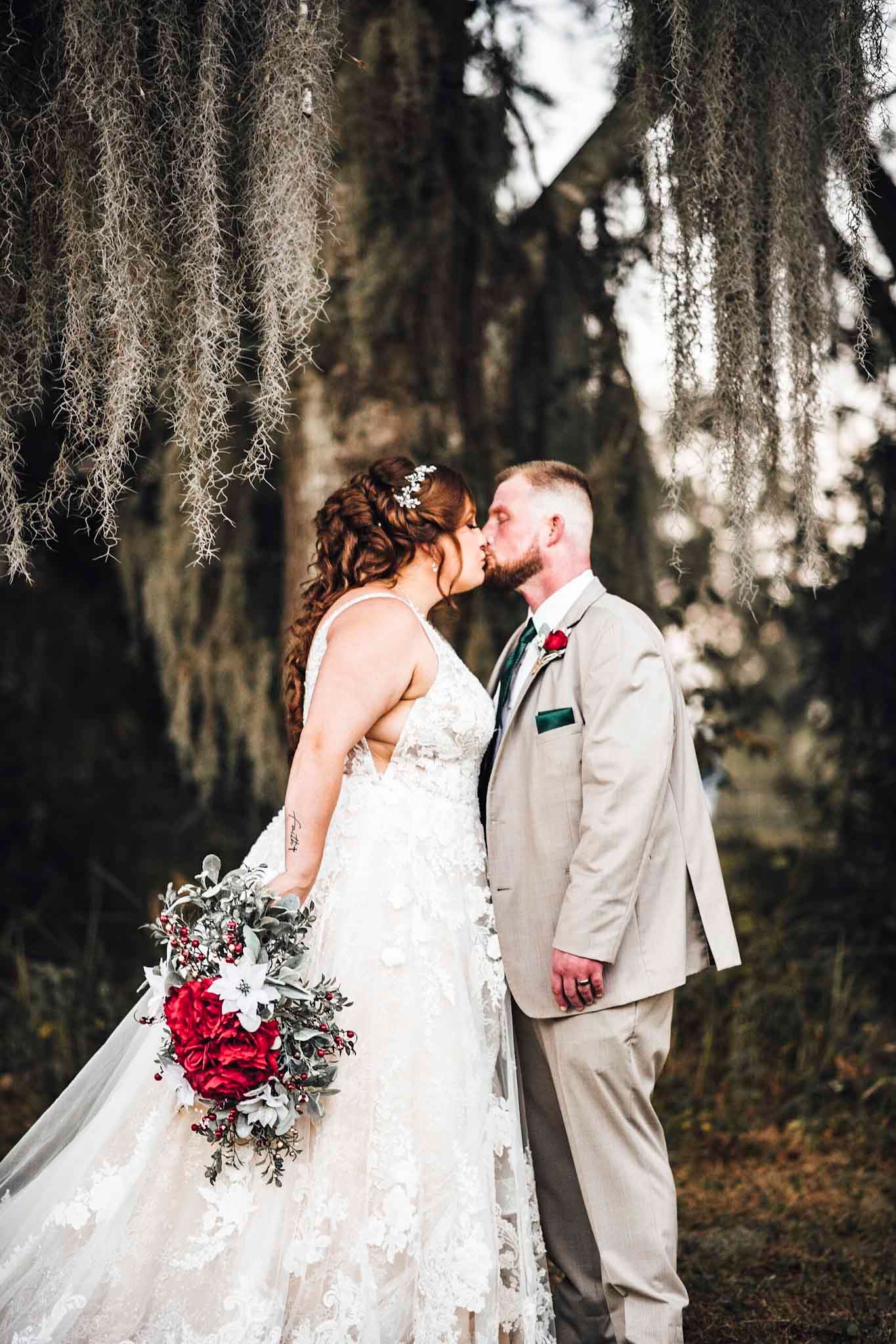 Wedding couple kissing outdoors under hanging Spanish moss, bride in lace dress with red floral bouquet, groom in beige suit.