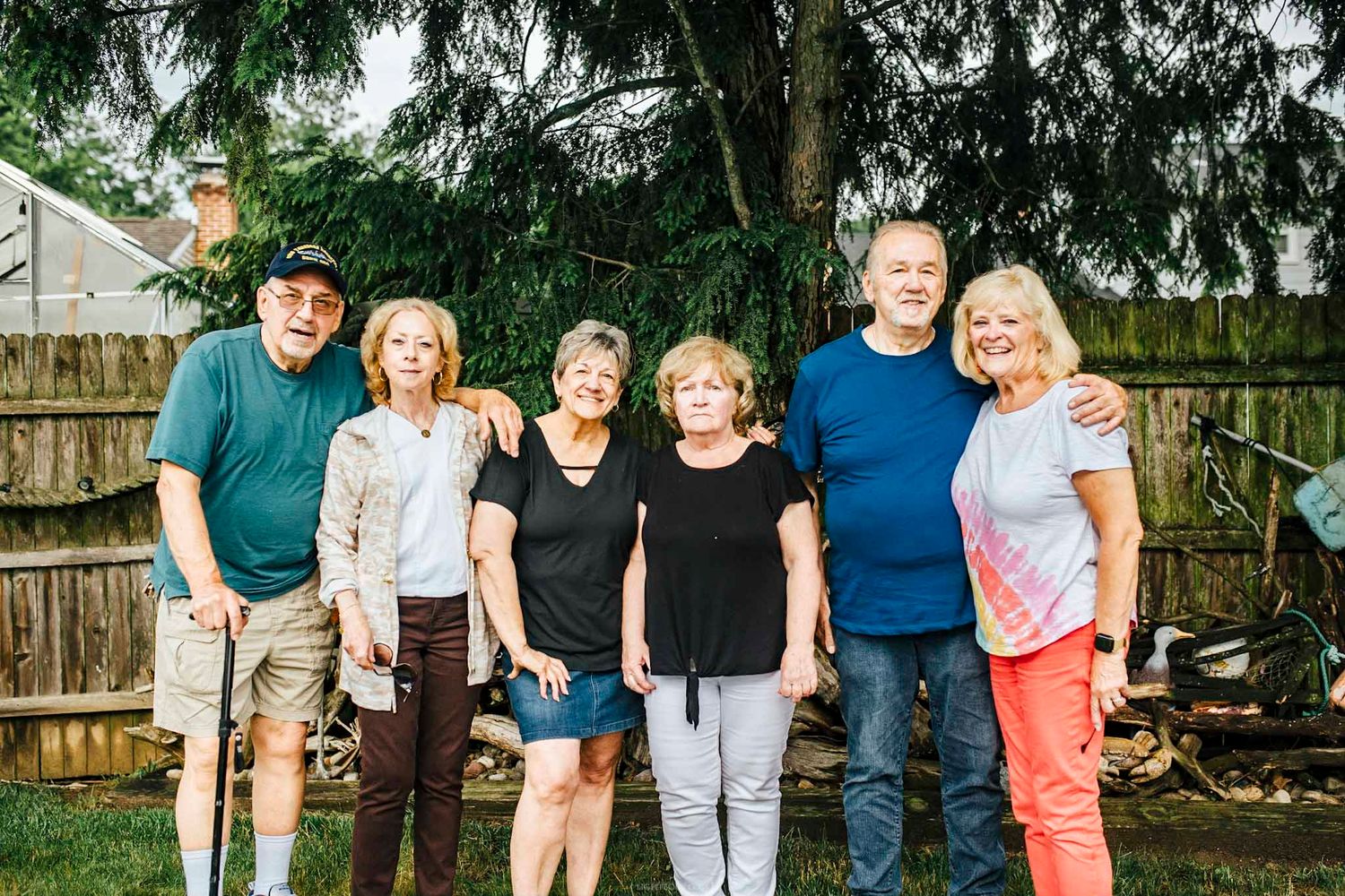 Six adults smiling together outdoors in a backyard, posing for a group photo near a wooden fence and evergreen tree.