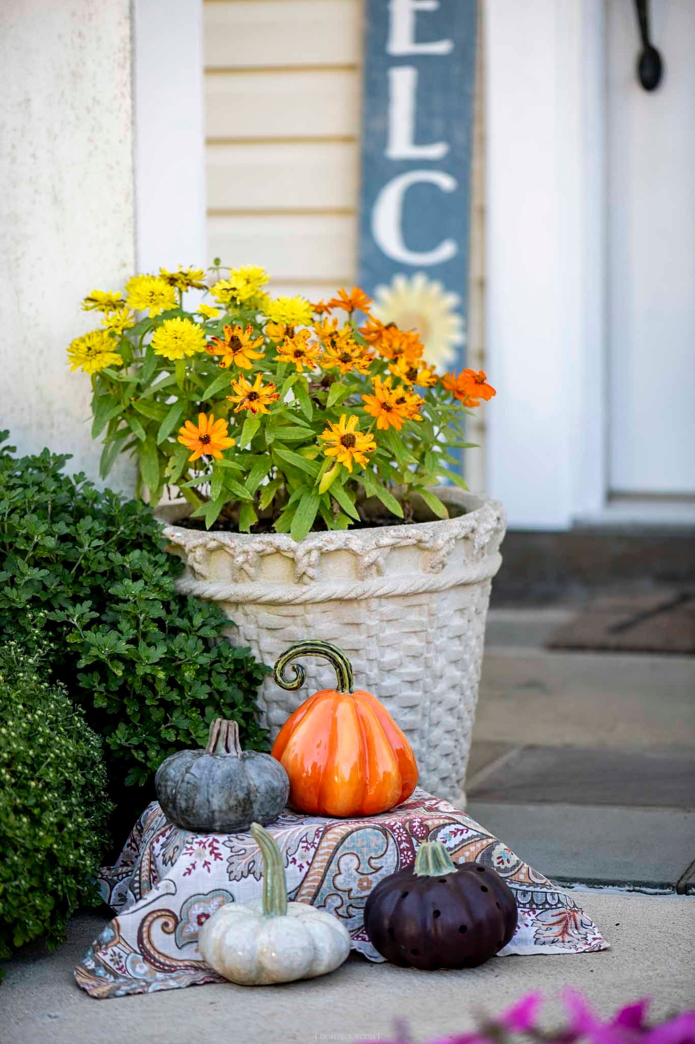 Fall porch decor with colorful pumpkins, paisley fabric, and yellow-orange flowers in a white basket planter near a welcome sign.