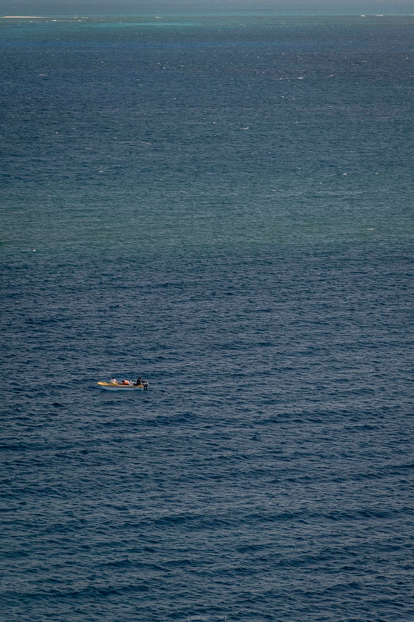 Lone small boat floating on calm deep blue ocean waters with a distant reef visible on the horizon.