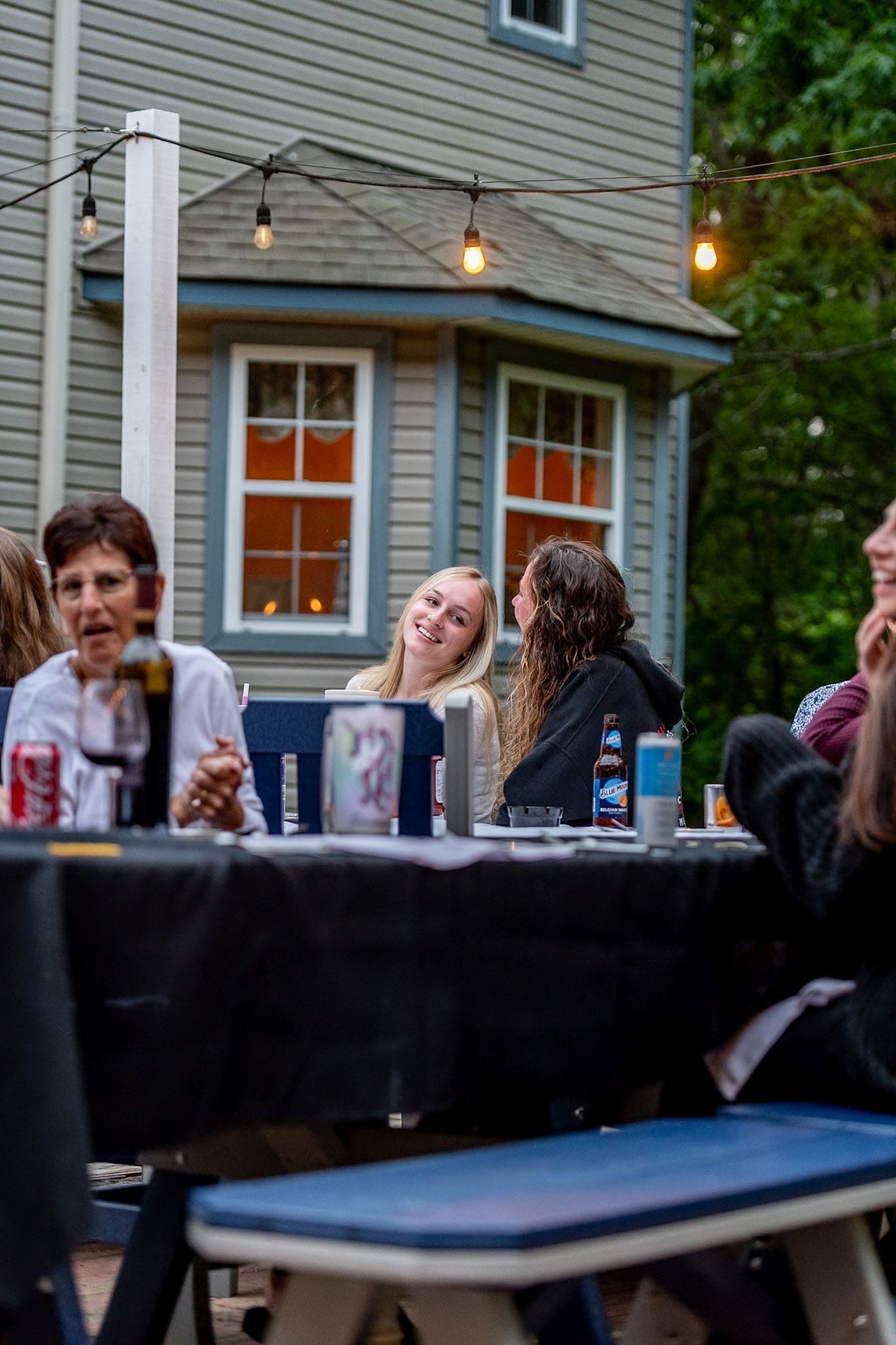 People socializing at long tables during an outdoor party with string lights and a gray house in the background.