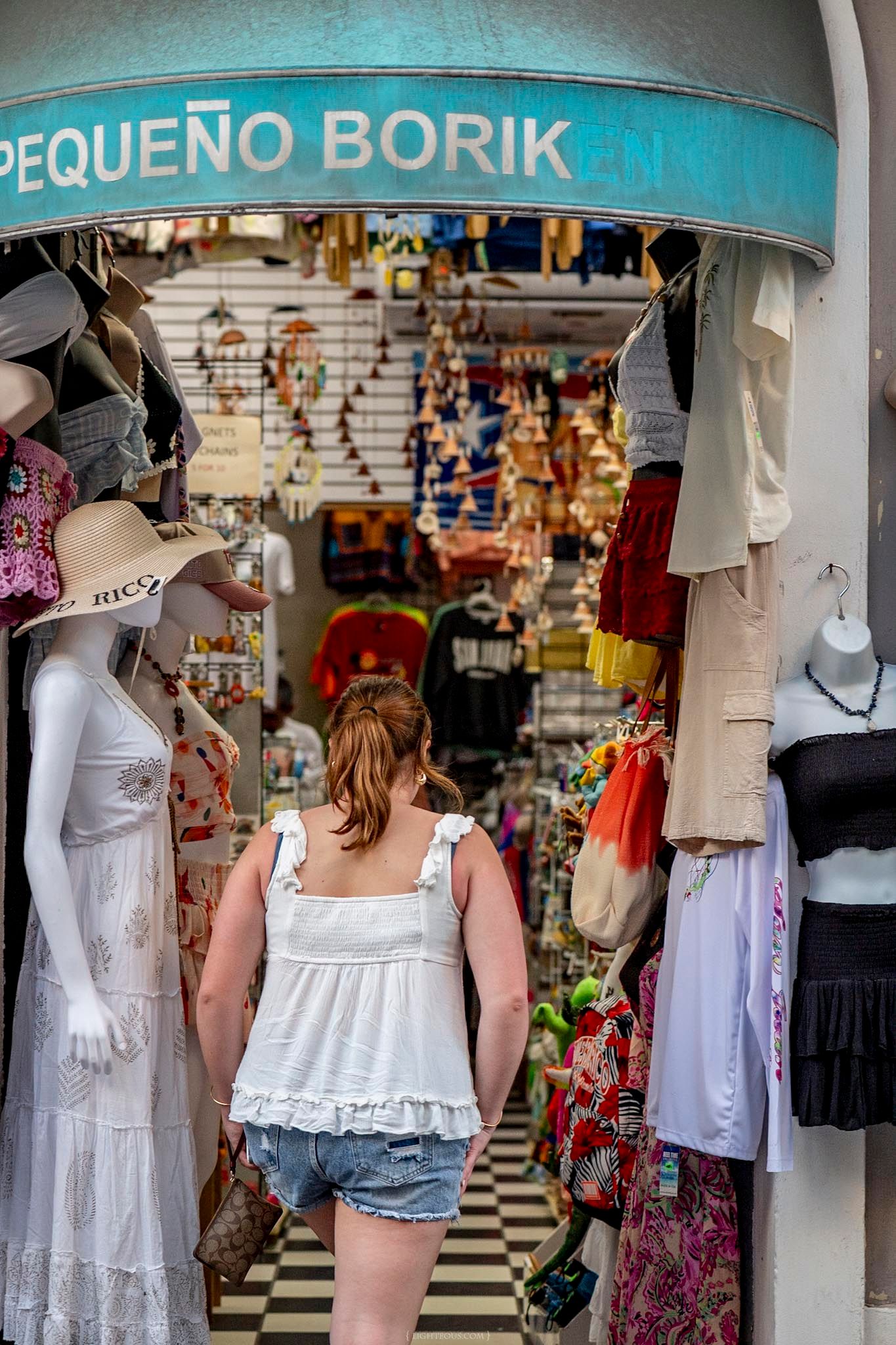 Young woman in white top and denim shorts browsing colorful clothing at Pequeno Borik shop entrance.