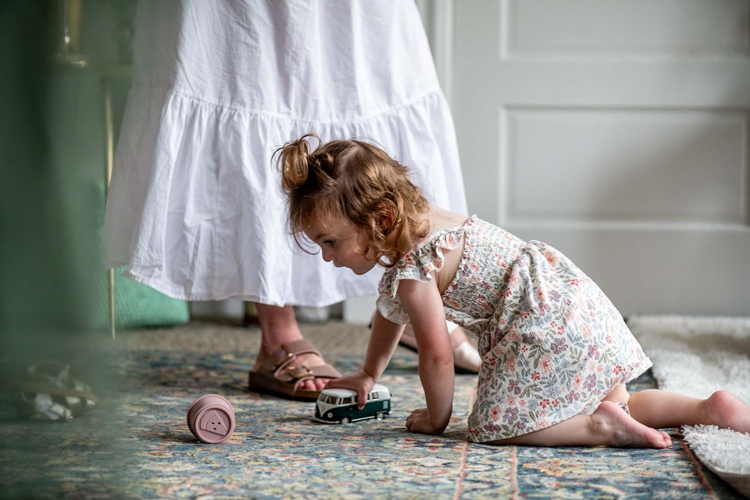 A white-dressed figure crawls playfully on a patterned carpet in a sunlit room with indoor plants.
