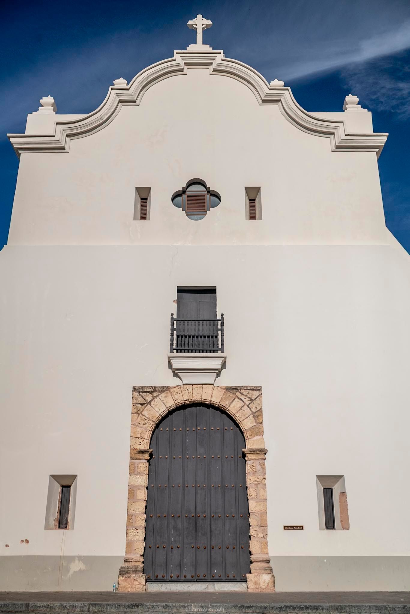 White Spanish colonial church facade with arched wooden door, cross atop ornate gable, and blue sky background.