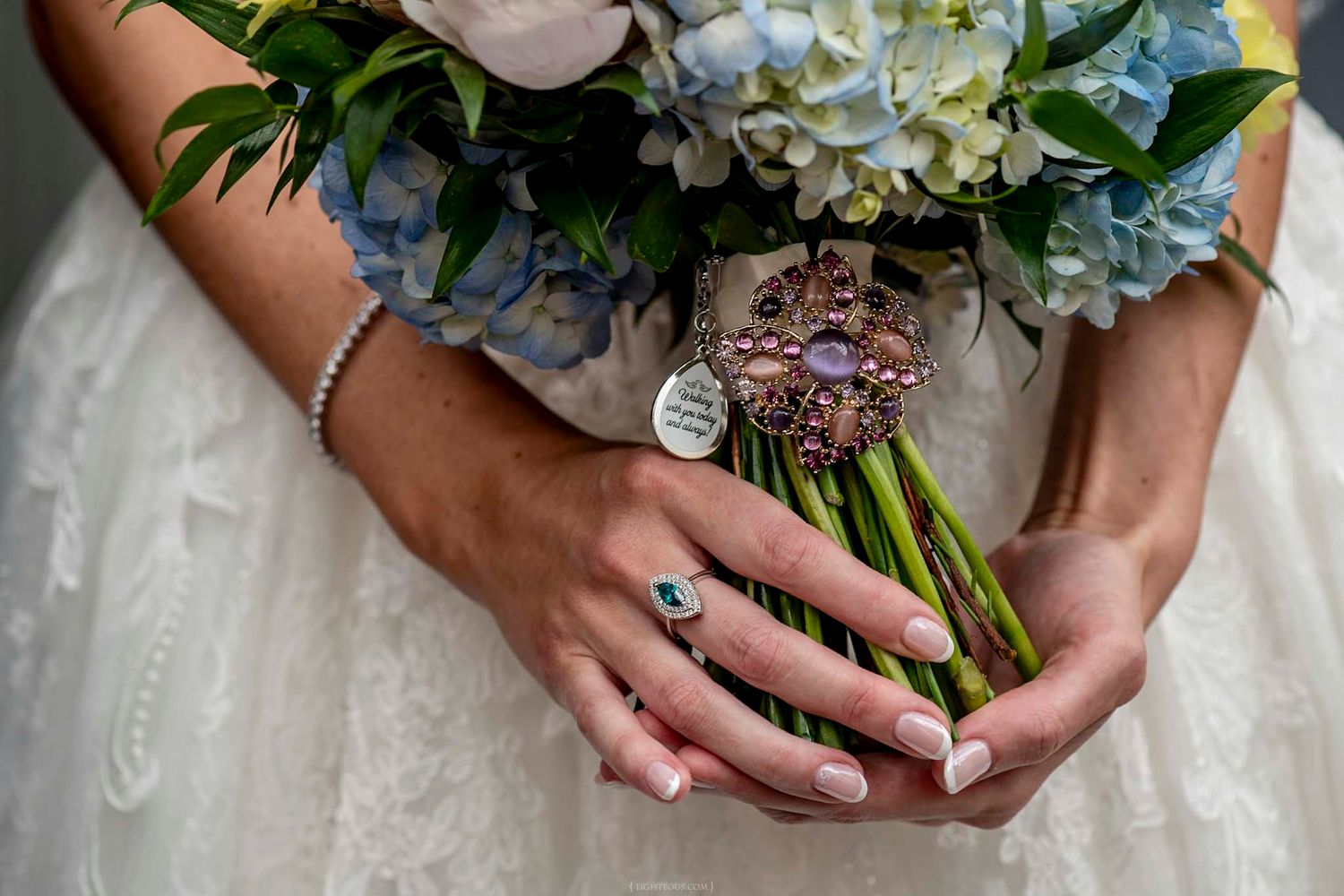 Bride holding blue hydrangea bouquet with brooch charm, wearing engagement ring and bracelet on wedding day.