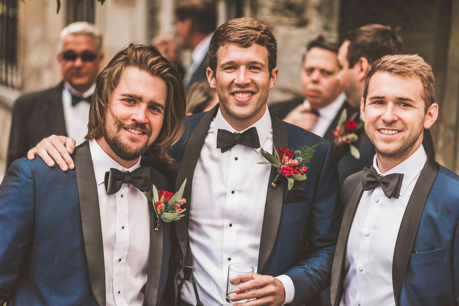 Three groomsmen in navy tuxedos with black bow ties and red boutonnieres smiling at a wedding celebration.