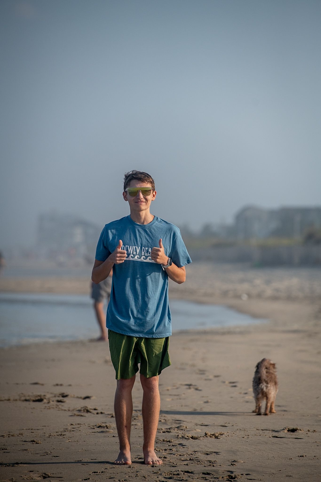 A young man in a blue t-shirt and green shorts gives thumbs up on a sandy beach with a dog nearby.