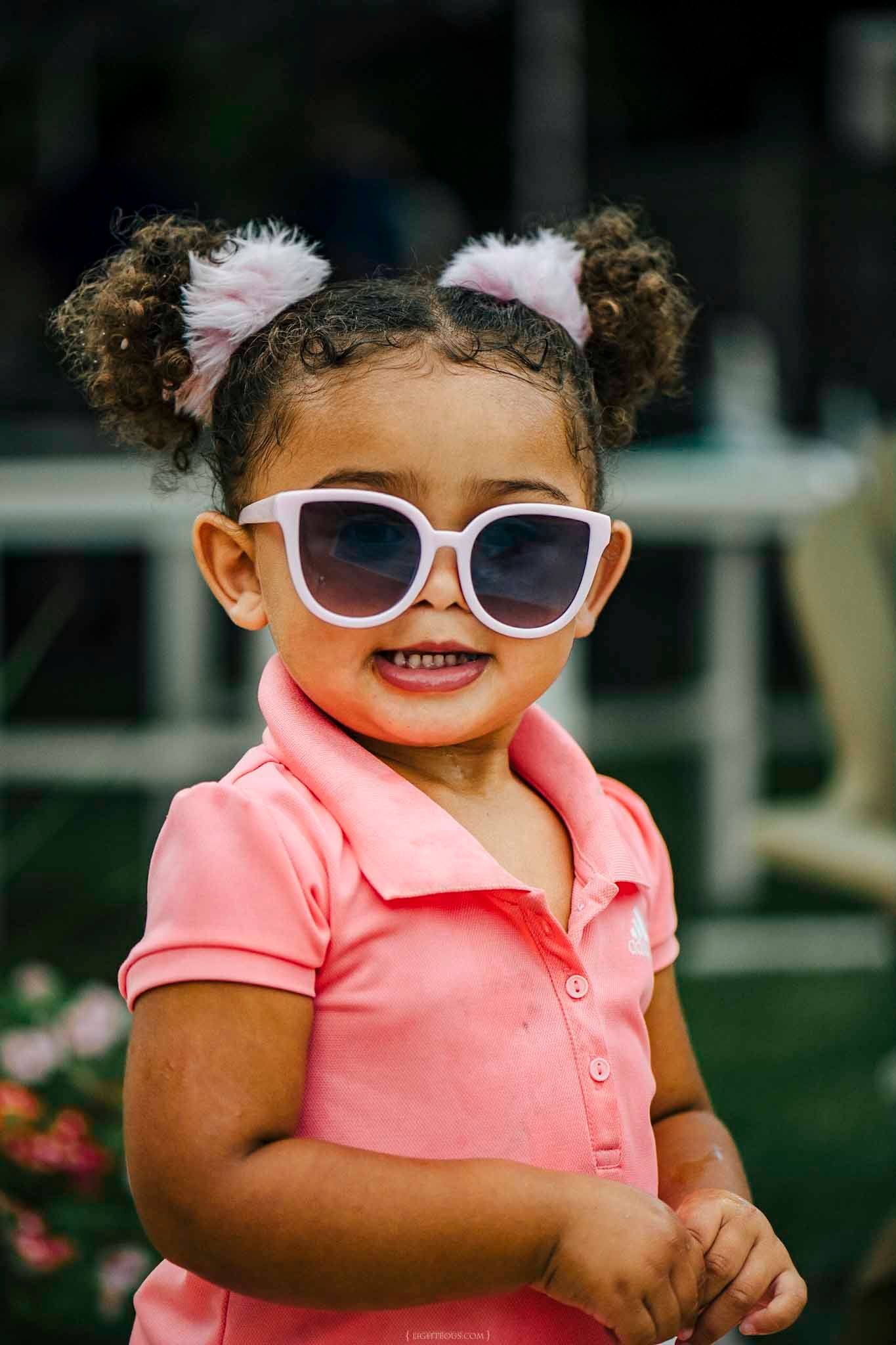 Adorable toddler girl wearing oversized white sunglasses and pink polo shirt with curly pigtails smiling outdoors.