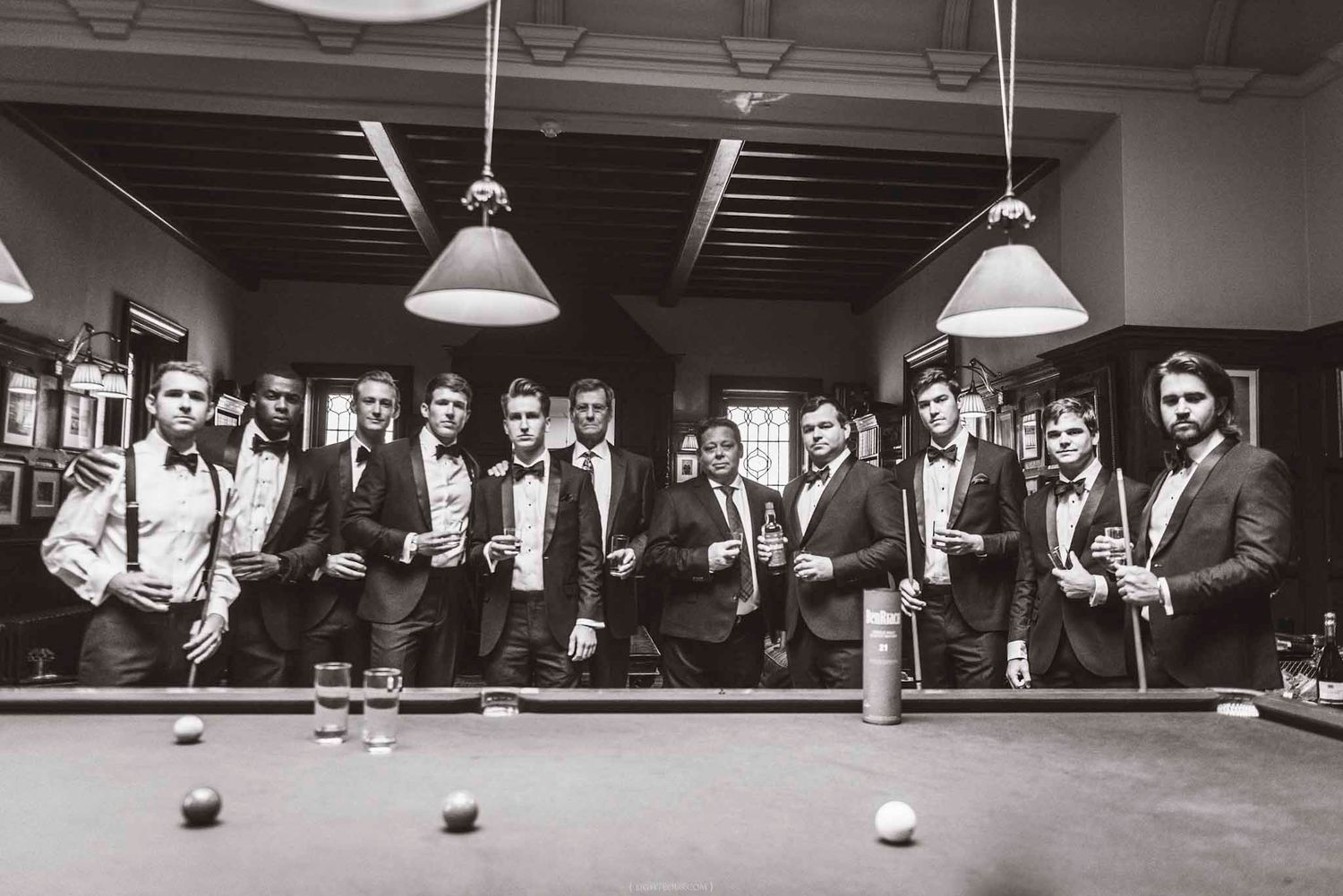 Black and white photo of groomsmen in tuxedos posing around a billiard table in an elegant venue.