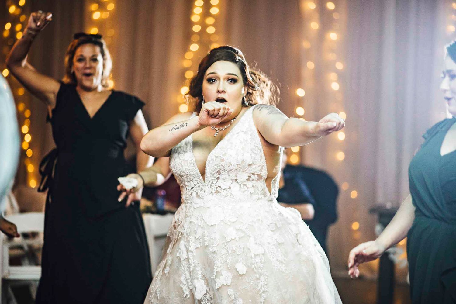 Bride in lace wedding gown blowing a kiss while dancing with guests at reception with warm fairy lights.