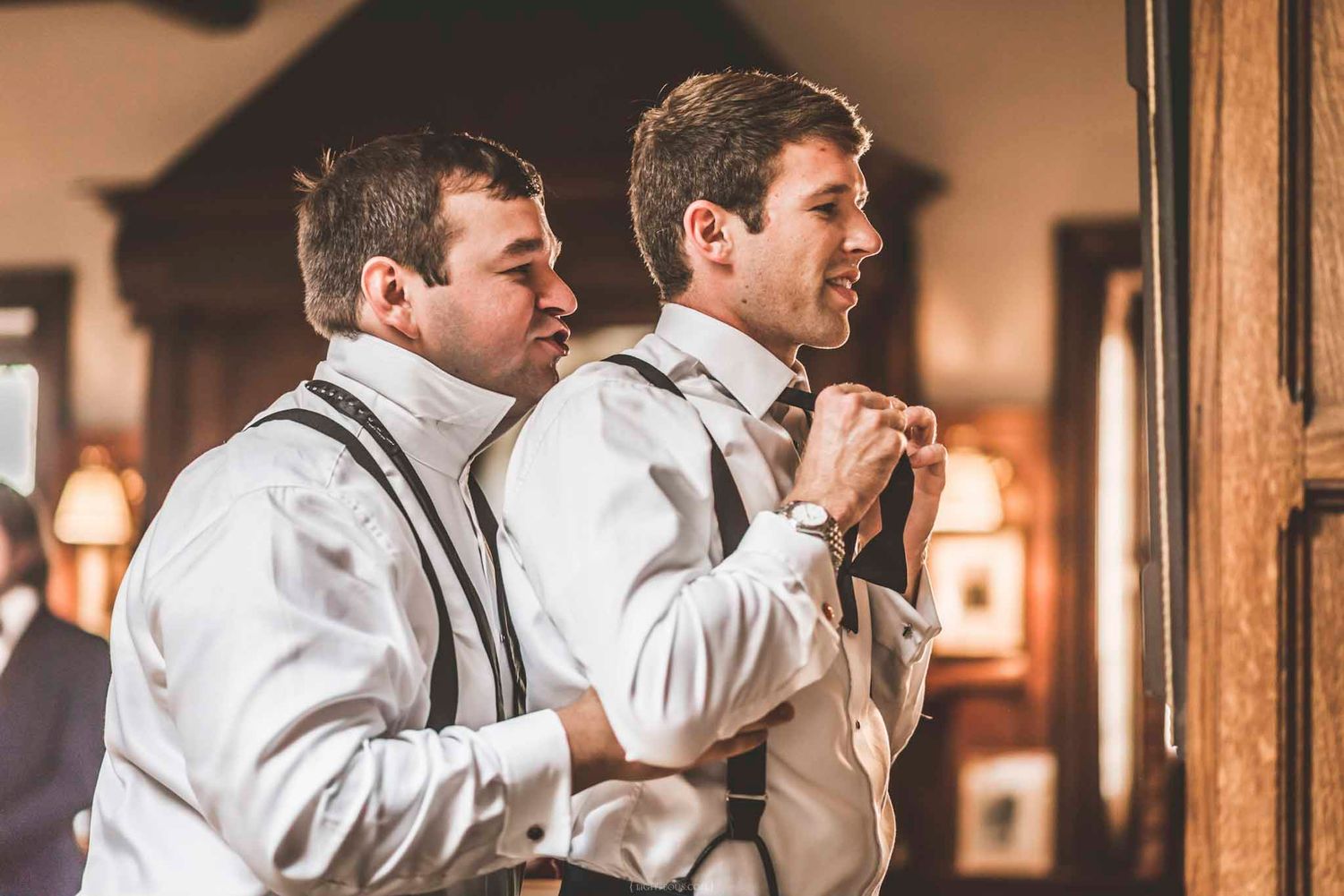 Two groomsmen in white shirts and suspenders adjusting neckties while preparing for a wedding ceremony.