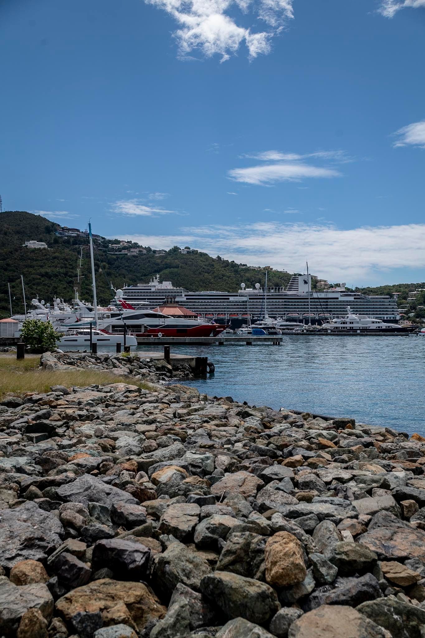 Rocky shoreline with cruise ships and yachts docked at a tropical harbor marina beneath a blue sky.