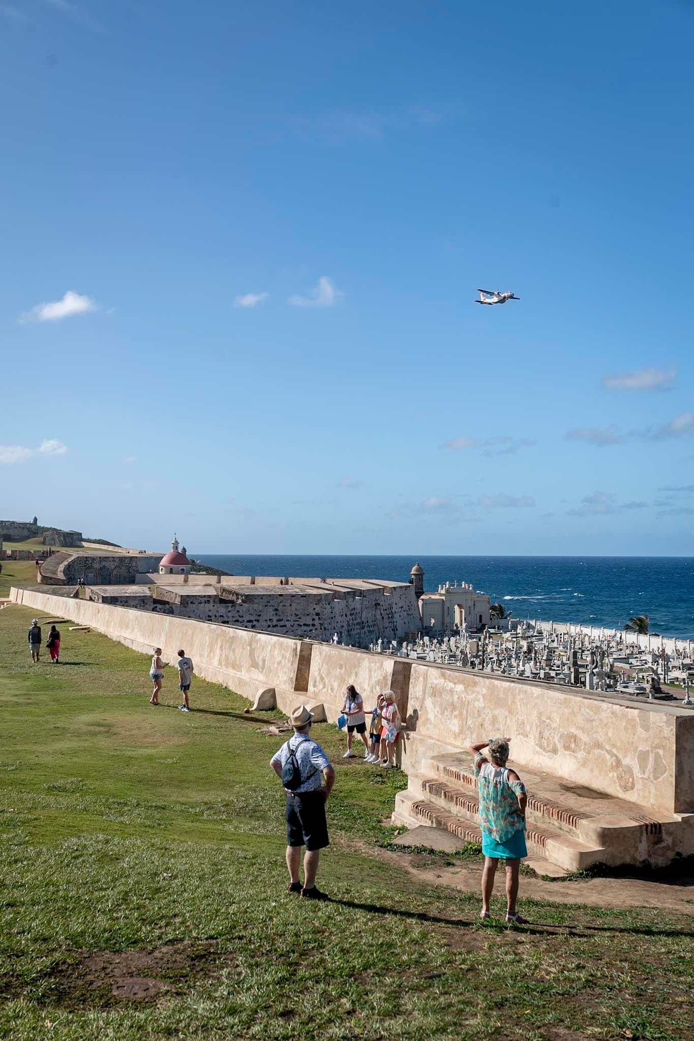 Tourists explore the historic walls of El Morro fort in San Juan, Puerto Rico, with a plane flying overhead and ocean views.