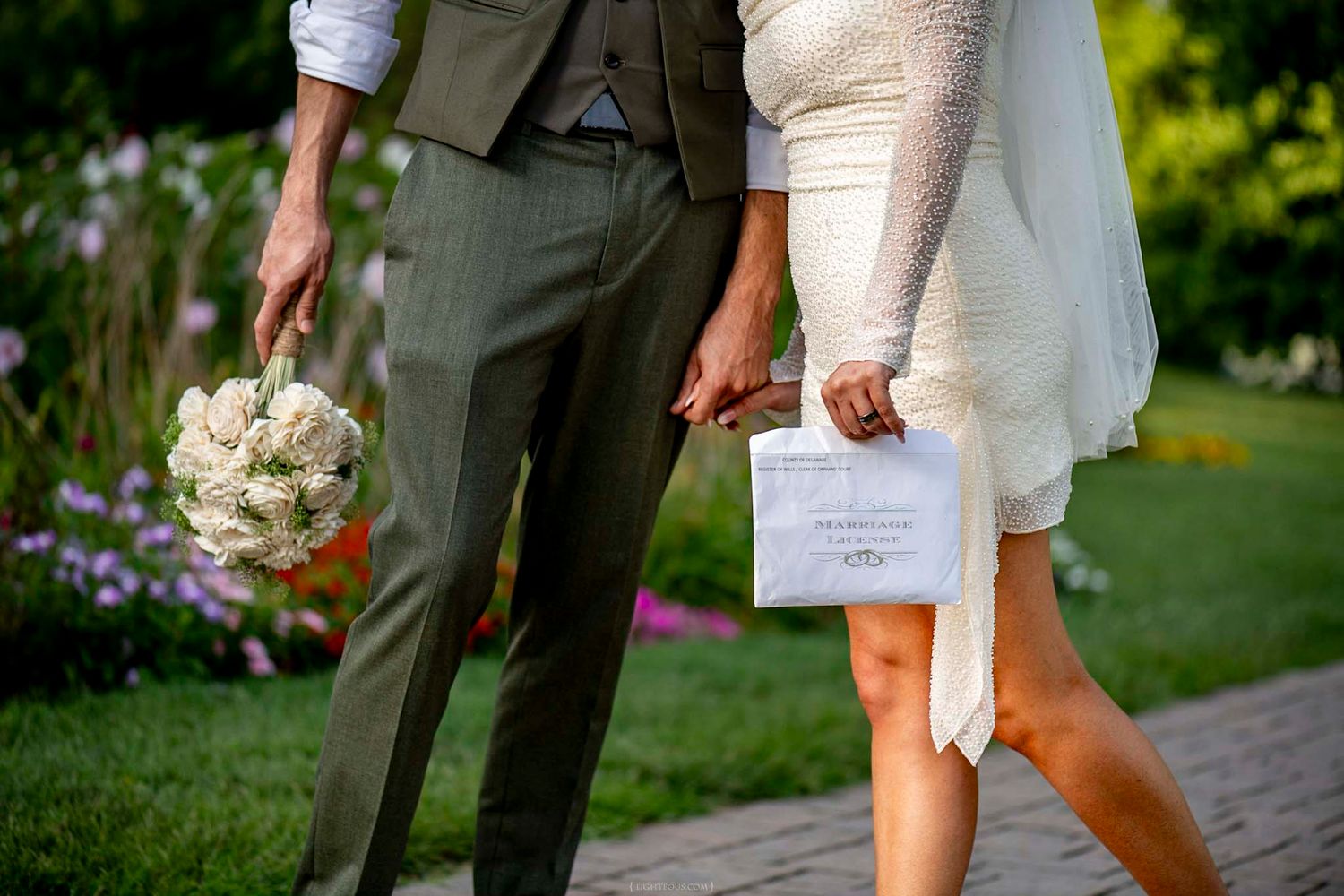 Bride and groom walking hand in hand, holding white bouquet and wedding program, in elegant outdoor garden setting.